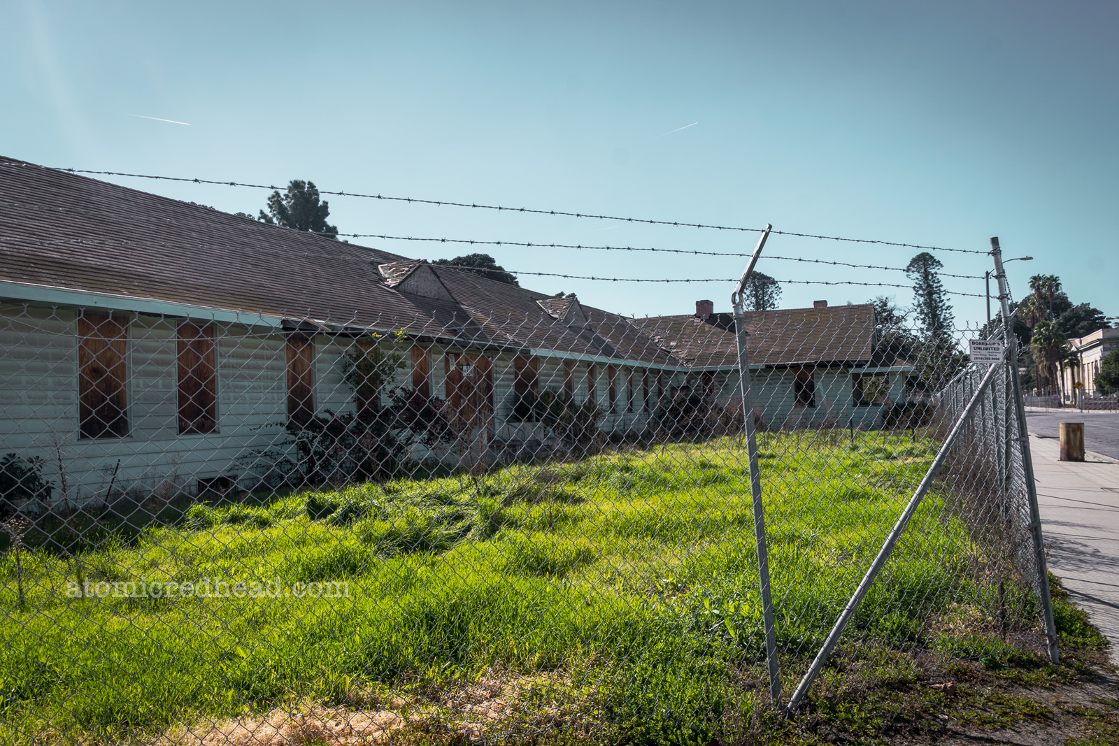 A boarded up single story building sits behind a chainlink fence with barbed wire atop.