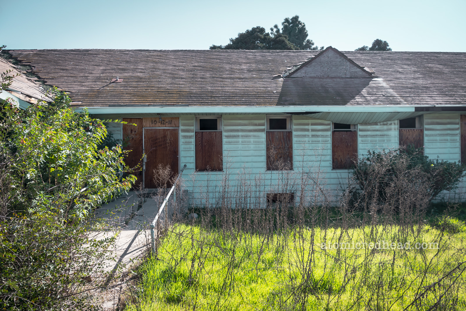A green yard with tall weeds sits in front of a long single story building with boarded up windows.