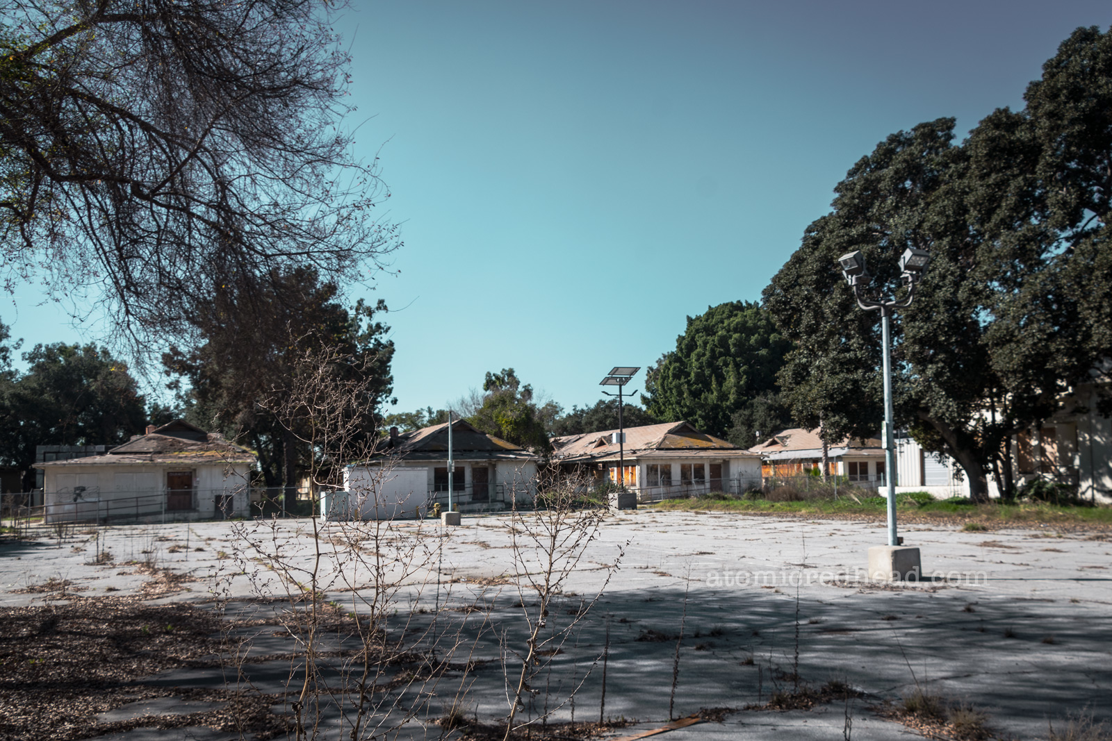 A slab of cracked pavement has weeds growing throughout it, as it stretches towards a cluster of single story wood buildings. All of which have been boarded up.