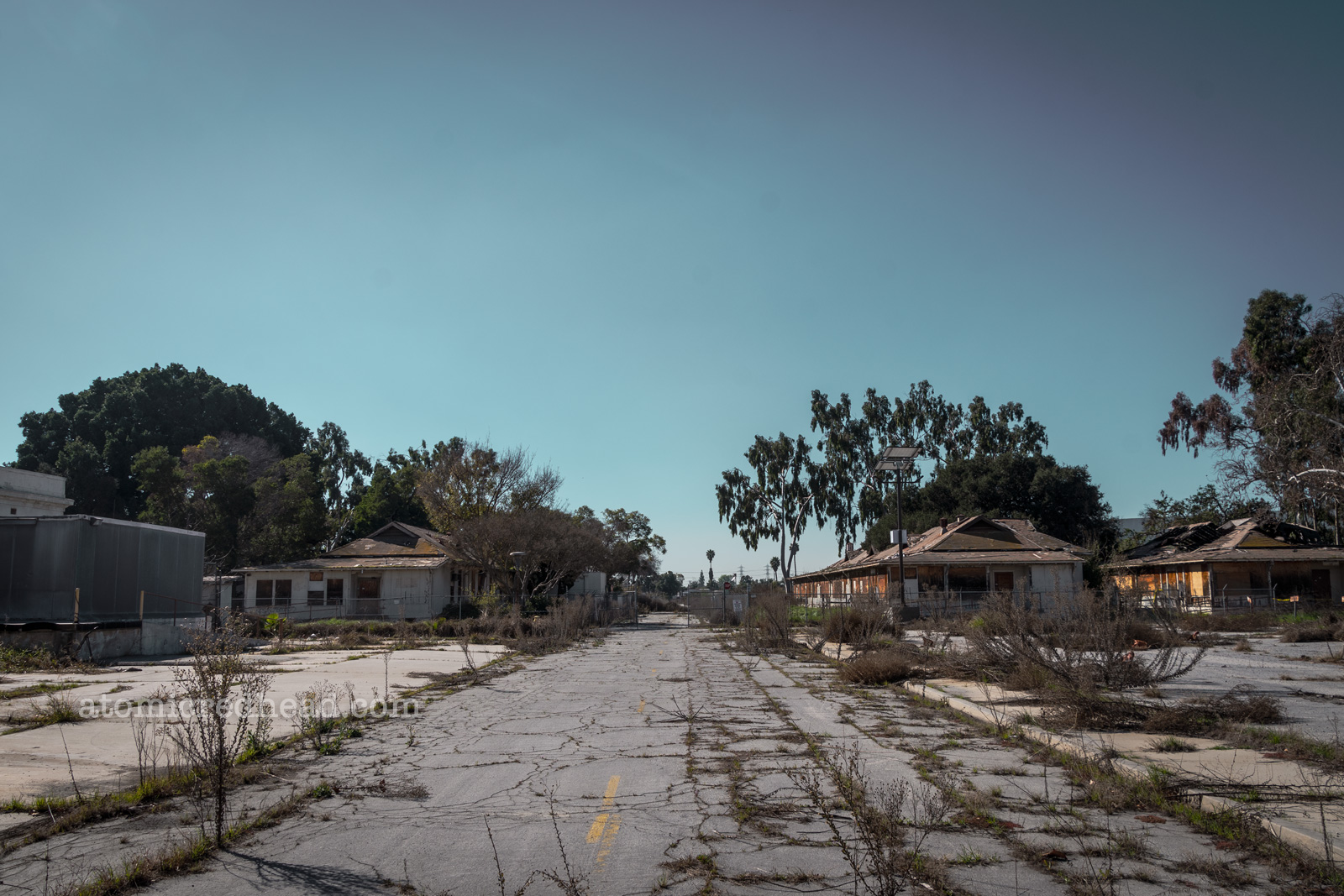 A cracked street with weeds growing through the cracks separates two single story wood structures. Each has been boarded up.