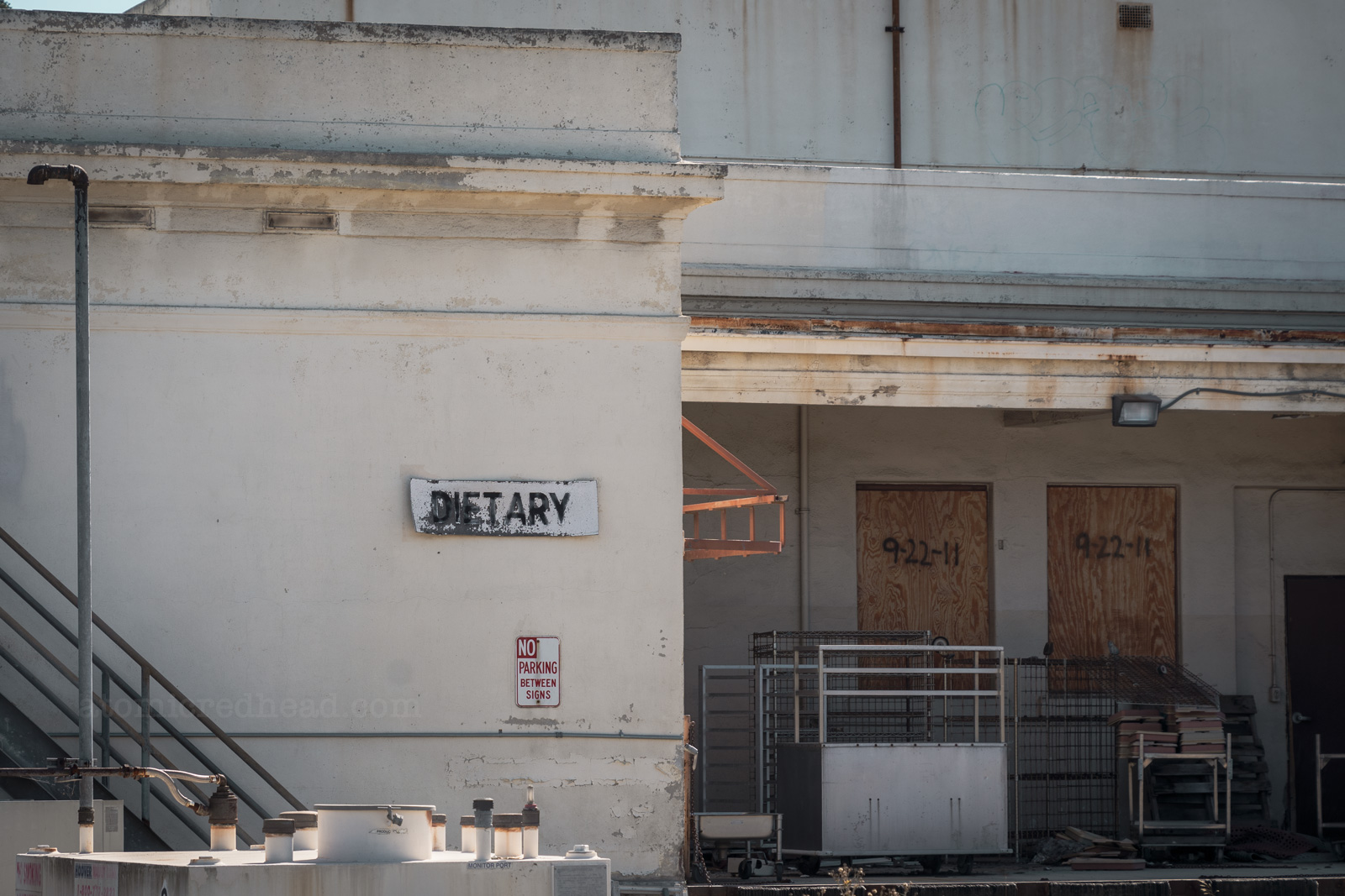 The docking bay of an industrial building. The doorways are boarded up. On the dock sits carts with food trays. A white sign reads "Dietary"