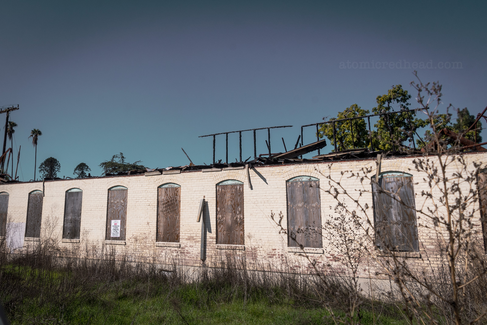 A long low building with boarded up windows shows signs of a fire. Charred pieces of wood still manage to stand.