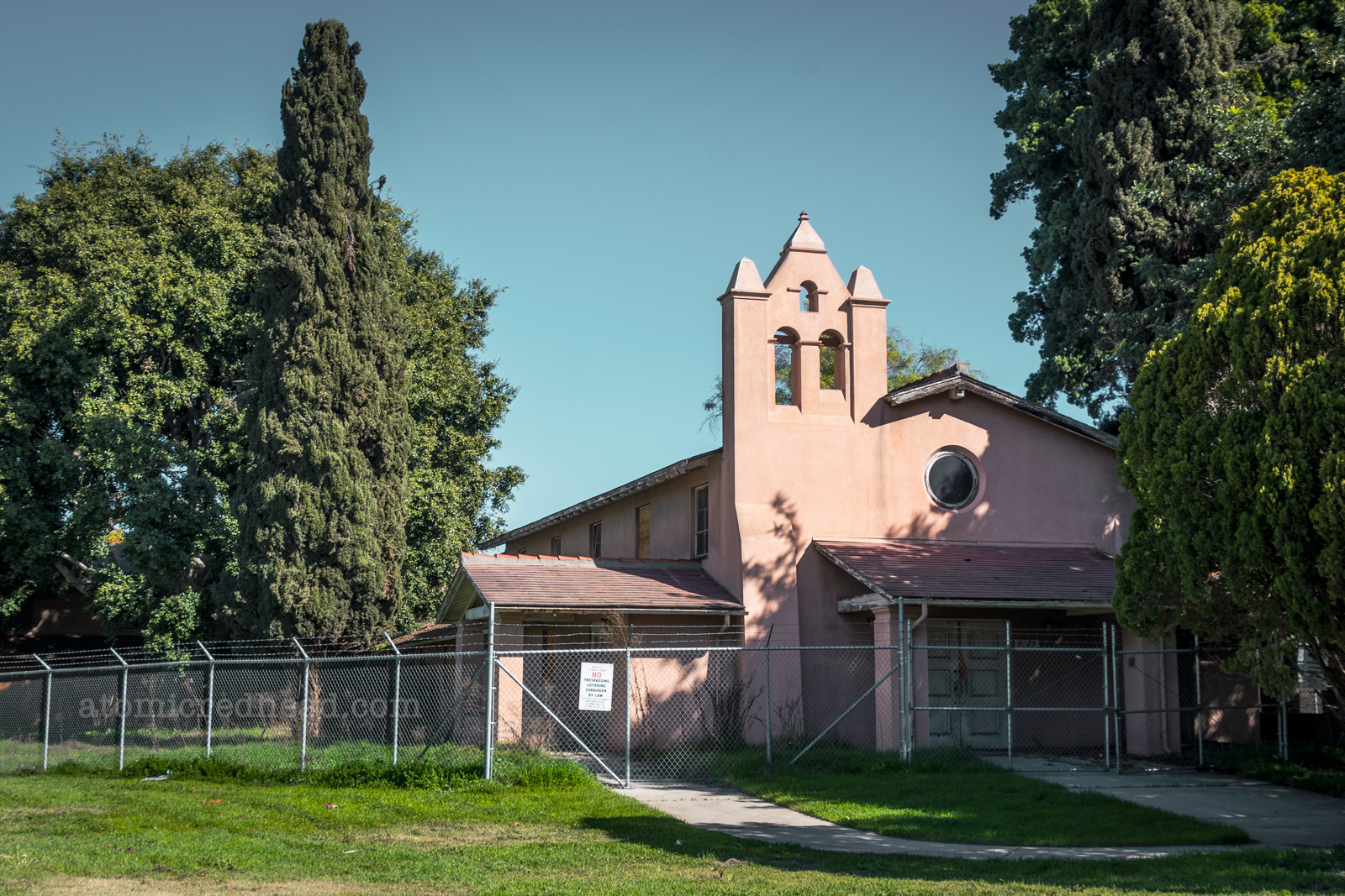A two story almost Spanish church like structure is painted a faded red color. A circular window is above a covered porch. A simple bell tower structure juts from the left side.