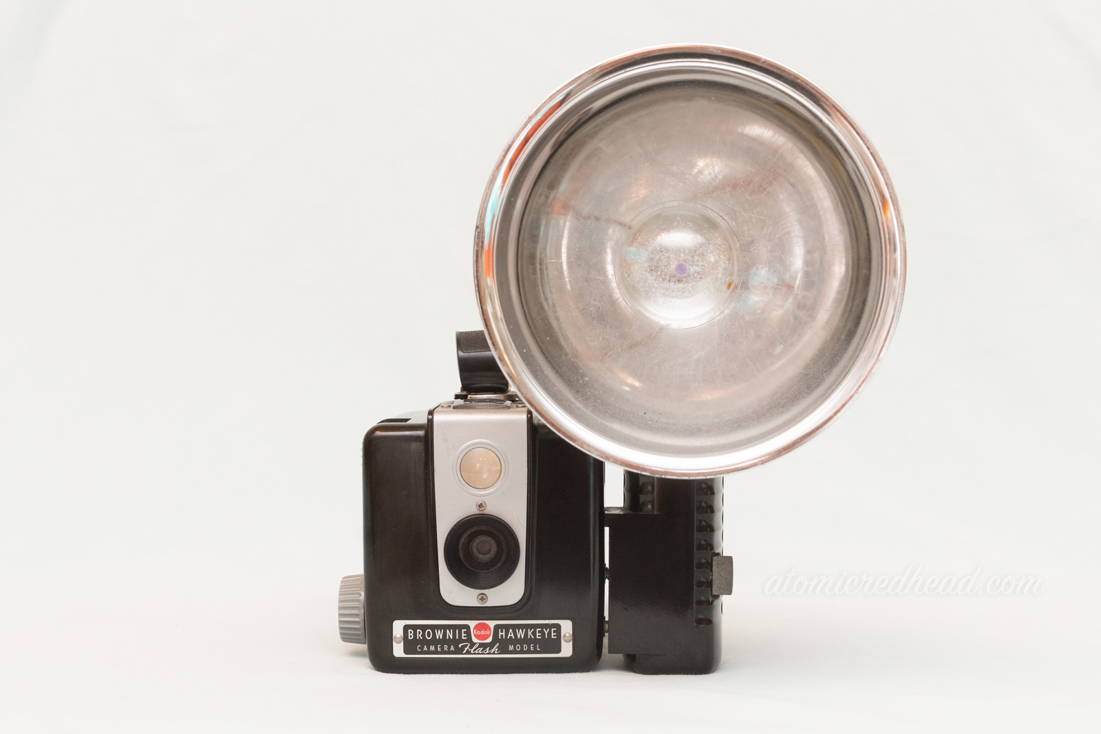 Kodak Brownie Hawkeye. A small square camera with a slim silver panel in the front around the lens. A flash attachment is on the right.