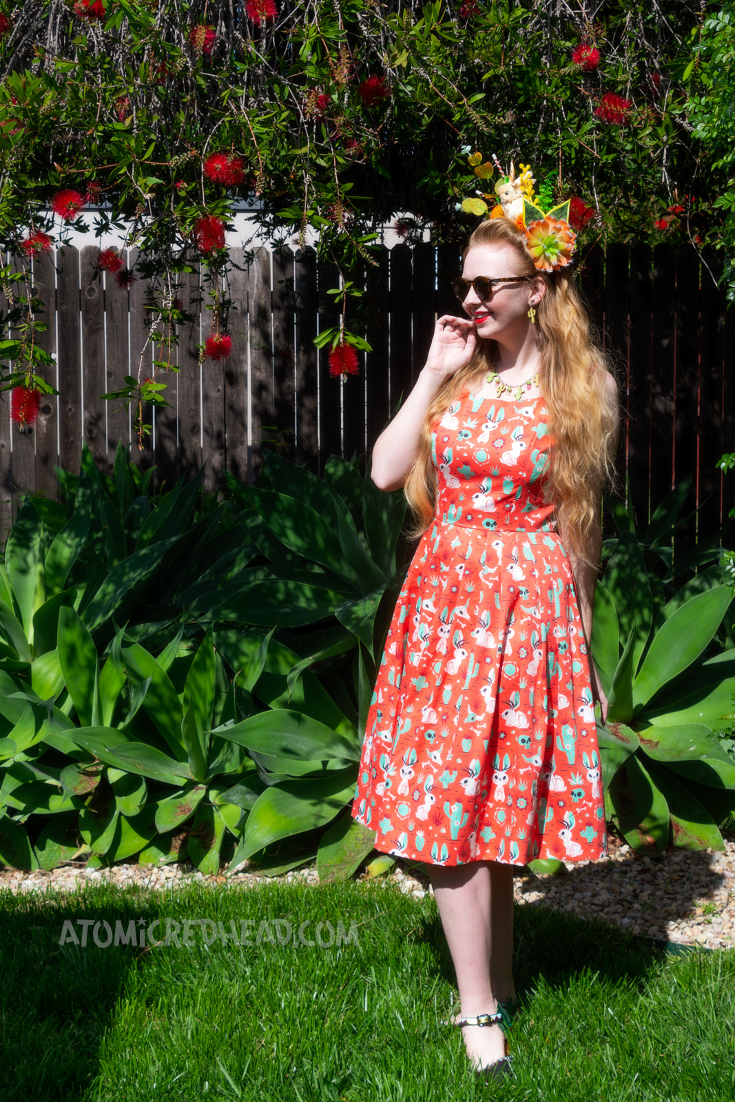 Myself wearing a crown made of fake succulents and a small cream jackalope, wearing a dress with a red, green, and cream print of cacti, jackalopes, and skulls, standing in our yard.