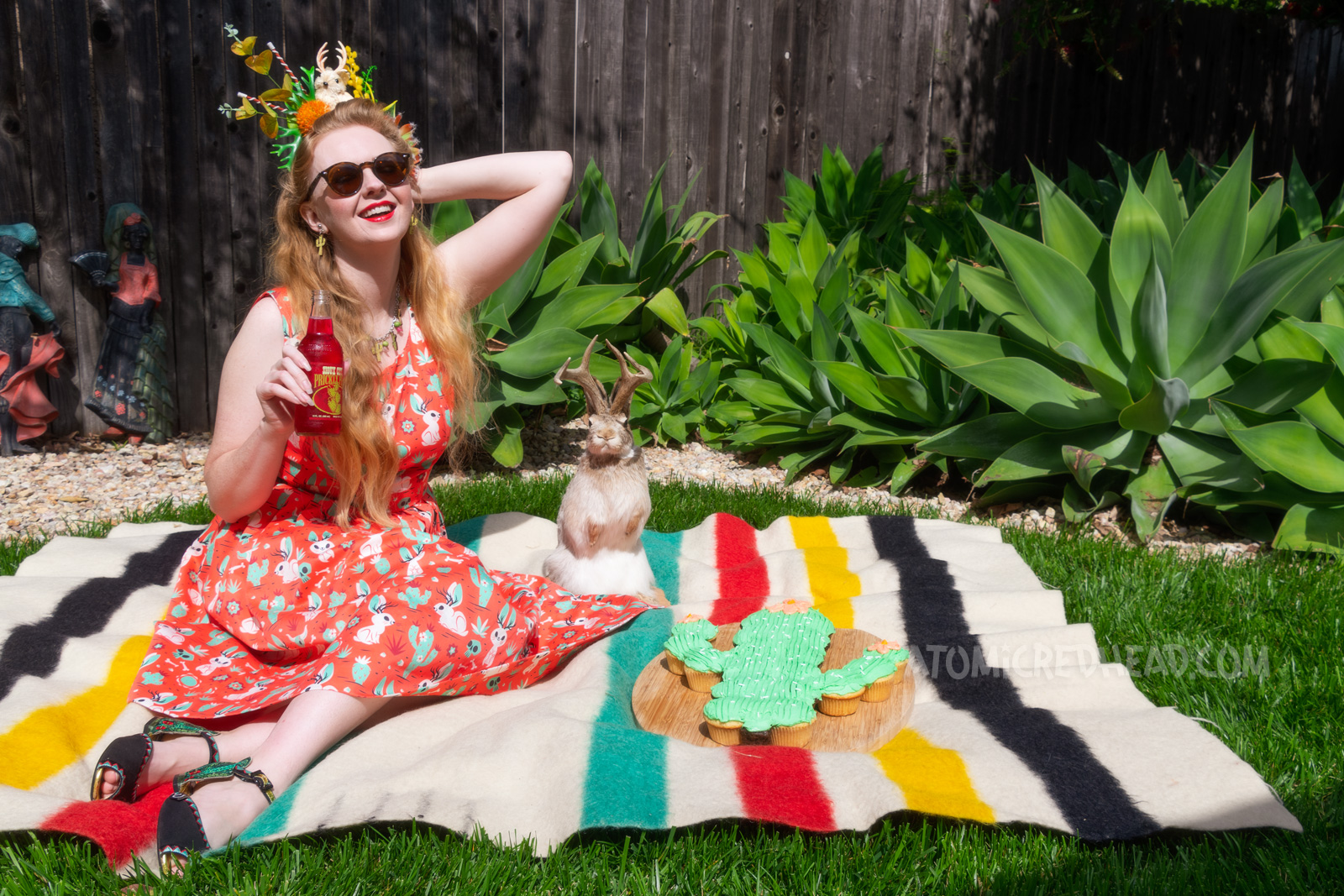 Myself wearing a crown made of fake succulents and a small cream jackalope, wearing a dress with a red, green, and cream print of cacti, jackalopes, and skulls, sitting on a blanket with a cactus shaped cake next to me, and a taxidermy jackalope nearby. 
