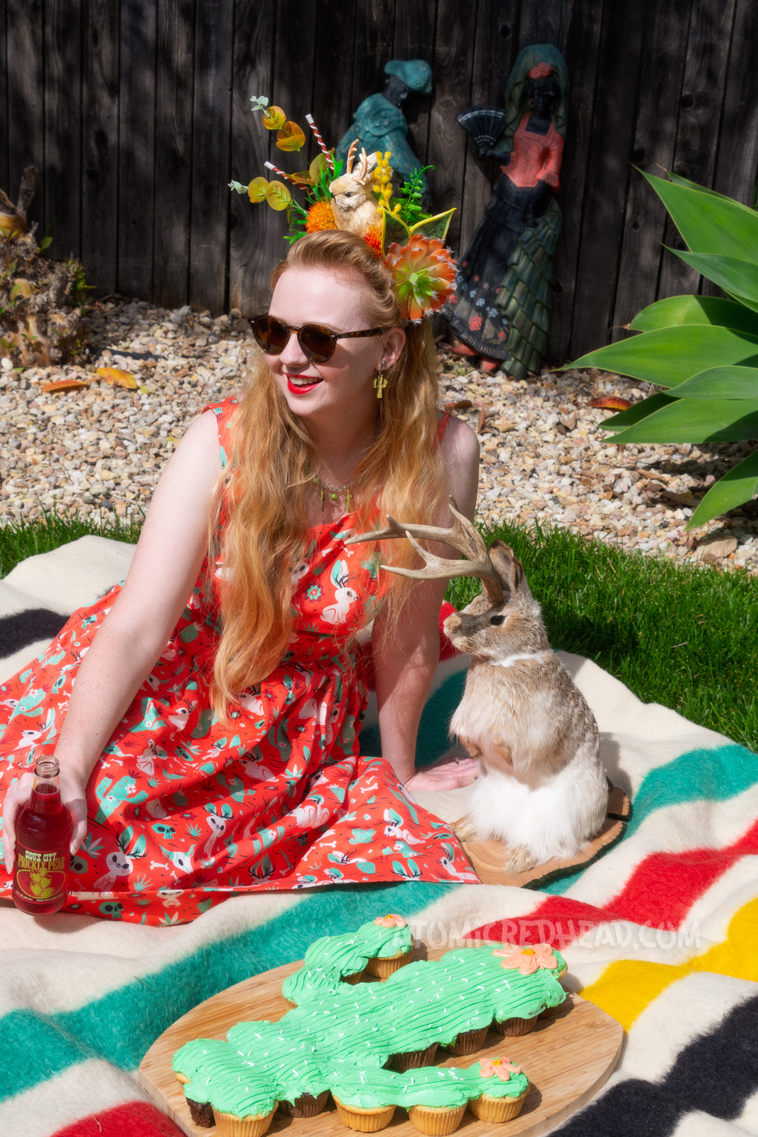 Myself wearing a crown made of fake succulents and a small cream jackalope, wearing a dress with a red, green, and cream print of cacti, jackalopes, and skulls, sitting on a blanket with a cactus shaped cake next to me, and a taxidermy jackalope nearby. 