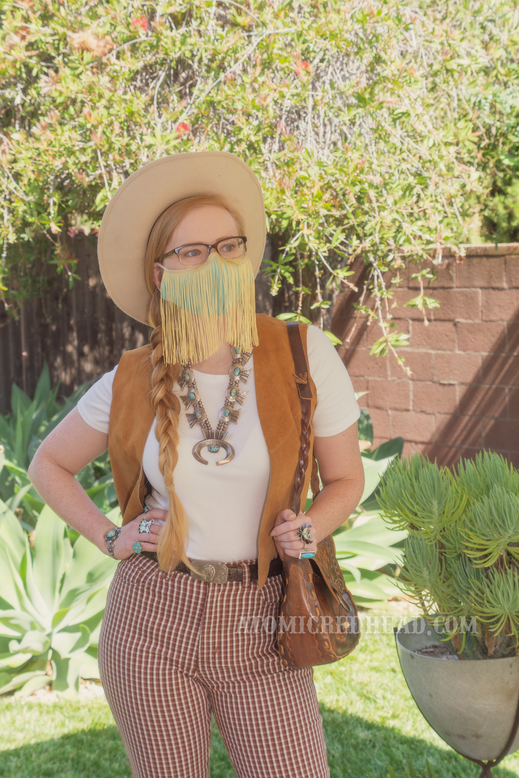 Myself, standing in my backyard, wearing a cream cowboy hat, a turquoise colored mask with gold fringe hanging from the top, a white t-shirt with a caramel colored suede vest over, a large turquoise squash blossom, and white and maroon plaid bell bottoms.