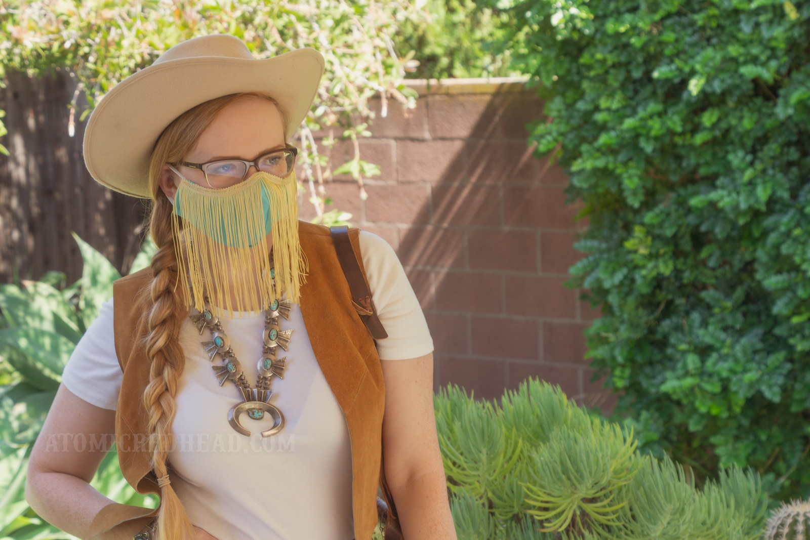 Myself, standing in my backyard, wearing a cream cowboy hat, a turquoise colored mask with gold fringe hanging from the top, a white t-shirt with a caramel colored suede vest over, a large turquoise squash blossom, and white and maroon plaid bell bottoms.
