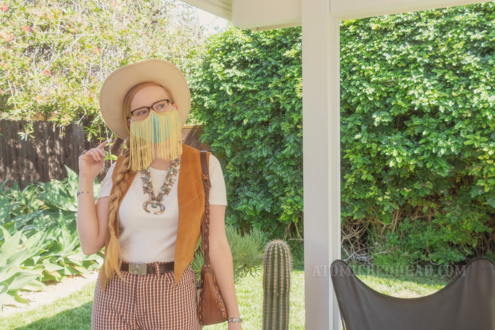 Myself, standing in my backyard, wearing a cream cowboy hat, a turquoise colored mask with gold fringe hanging from the top, a white t-shirt with a caramel colored suede vest over, a large turquoise squash blossom, and white and maroon plaid bell bottoms.