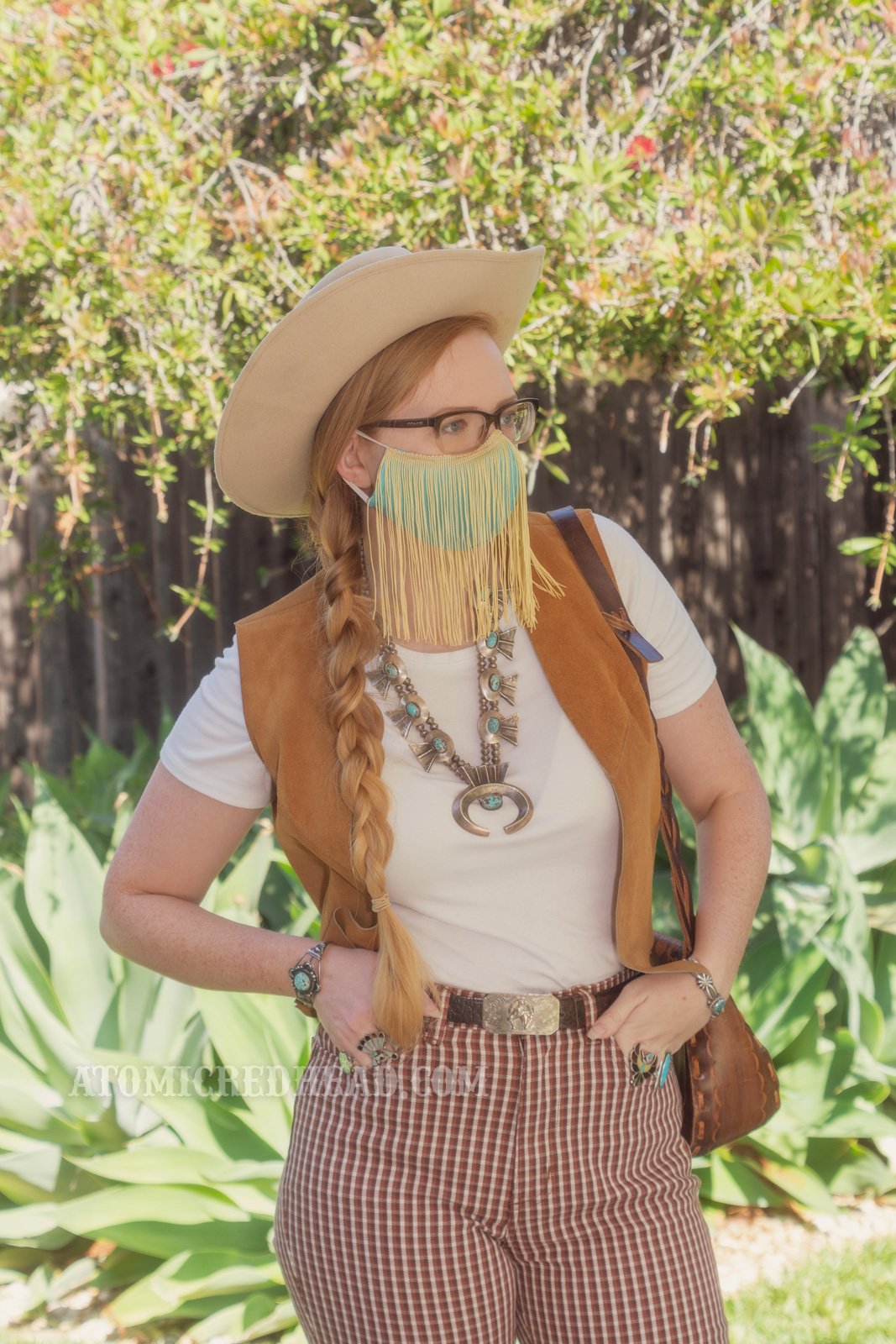 Myself, standing in my backyard, wearing a cream cowboy hat, a turquoise colored mask with gold fringe hanging from the top, a white t-shirt with a caramel colored suede vest over, a large turquoise squash blossom, and white and maroon plaid bell bottoms.