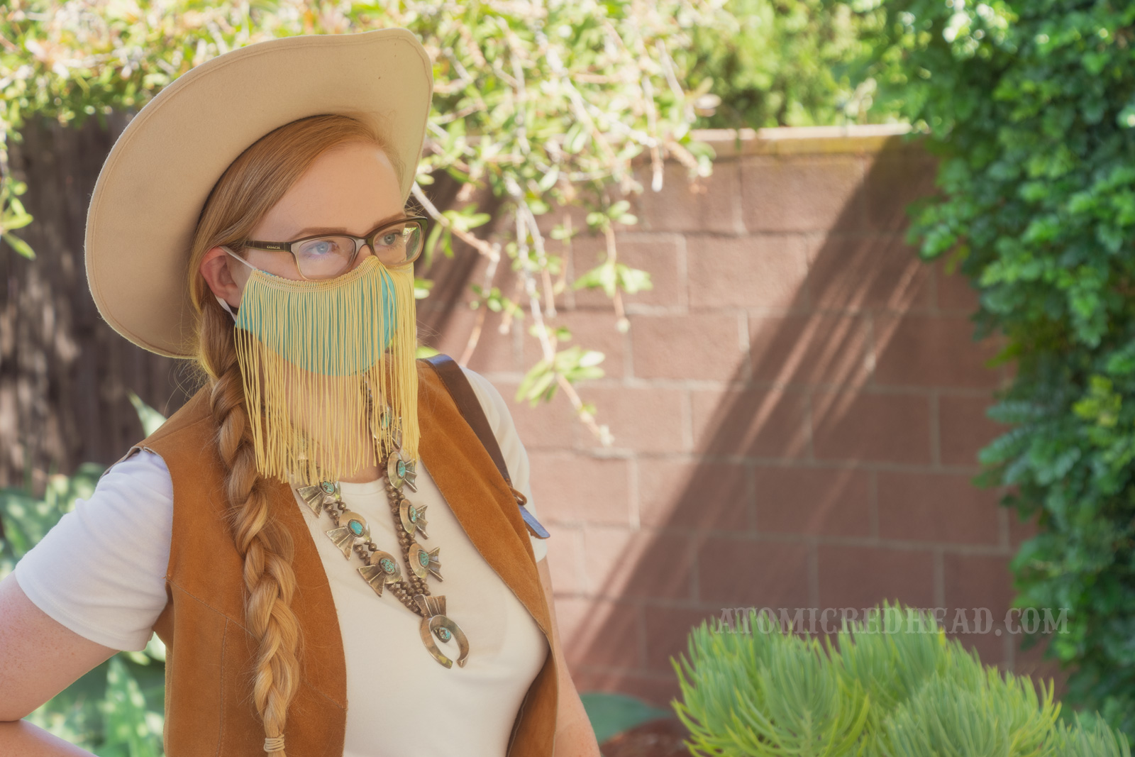 Myself, standing in my backyard, wearing a cream cowboy hat, a turquoise colored mask with gold fringe hanging from the top, a white t-shirt with a caramel colored suede vest over, a large turquoise squash blossom, and white and maroon plaid bell bottoms.