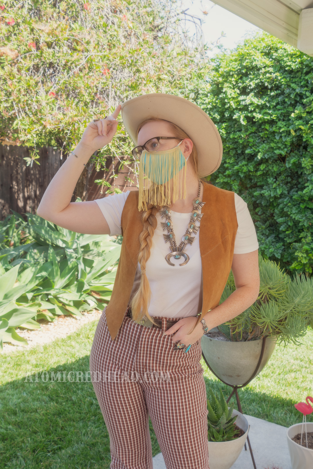 Myself, standing in my backyard, wearing a cream cowboy hat, a turquoise colored mask with gold fringe hanging from the top, a white t-shirt with a caramel colored suede vest over, a large turquoise squash blossom, and white and maroon plaid bell bottoms.