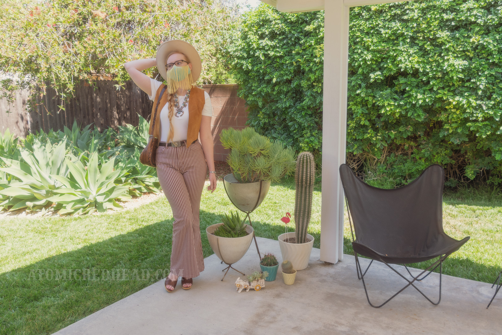 Myself, standing in my backyard, wearing a cream cowboy hat, a turquoise colored mask with gold fringe hanging from the top, a white t-shirt with a caramel colored suede vest over, a large turquoise squash blossom, and white and maroon plaid bell bottoms. Various planted cacti are visible on the right.