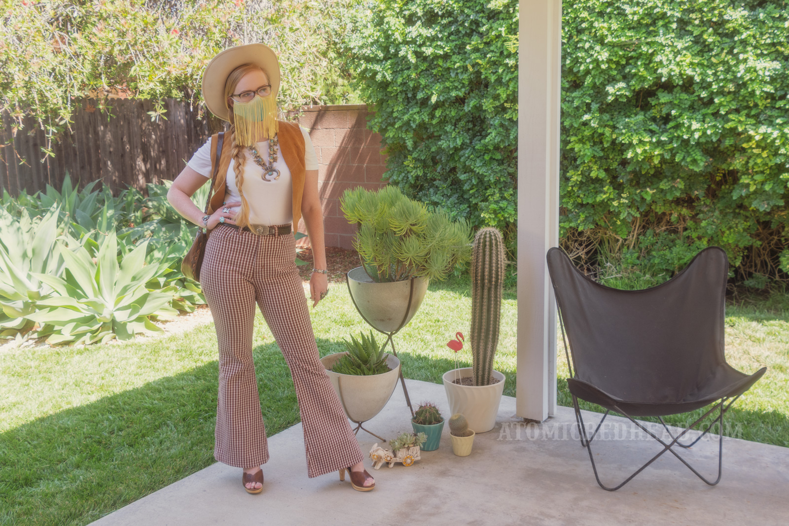 Myself, standing in my backyard, wearing a cream cowboy hat, a turquoise colored mask with gold fringe hanging from the top, a white t-shirt with a caramel colored suede vest over, a large turquoise squash blossom, and white and maroon plaid bell bottoms. Various planted cacti are visible on the right.