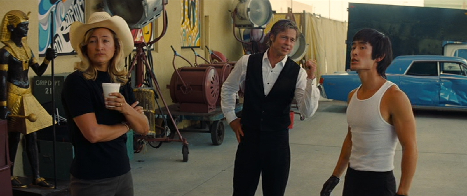 Janet stands with her arms crossed next to Cliff, a man in a white button up shirt, black vest, and black pants, and Bruce Lee, a man wearing a white tank top and black pants.