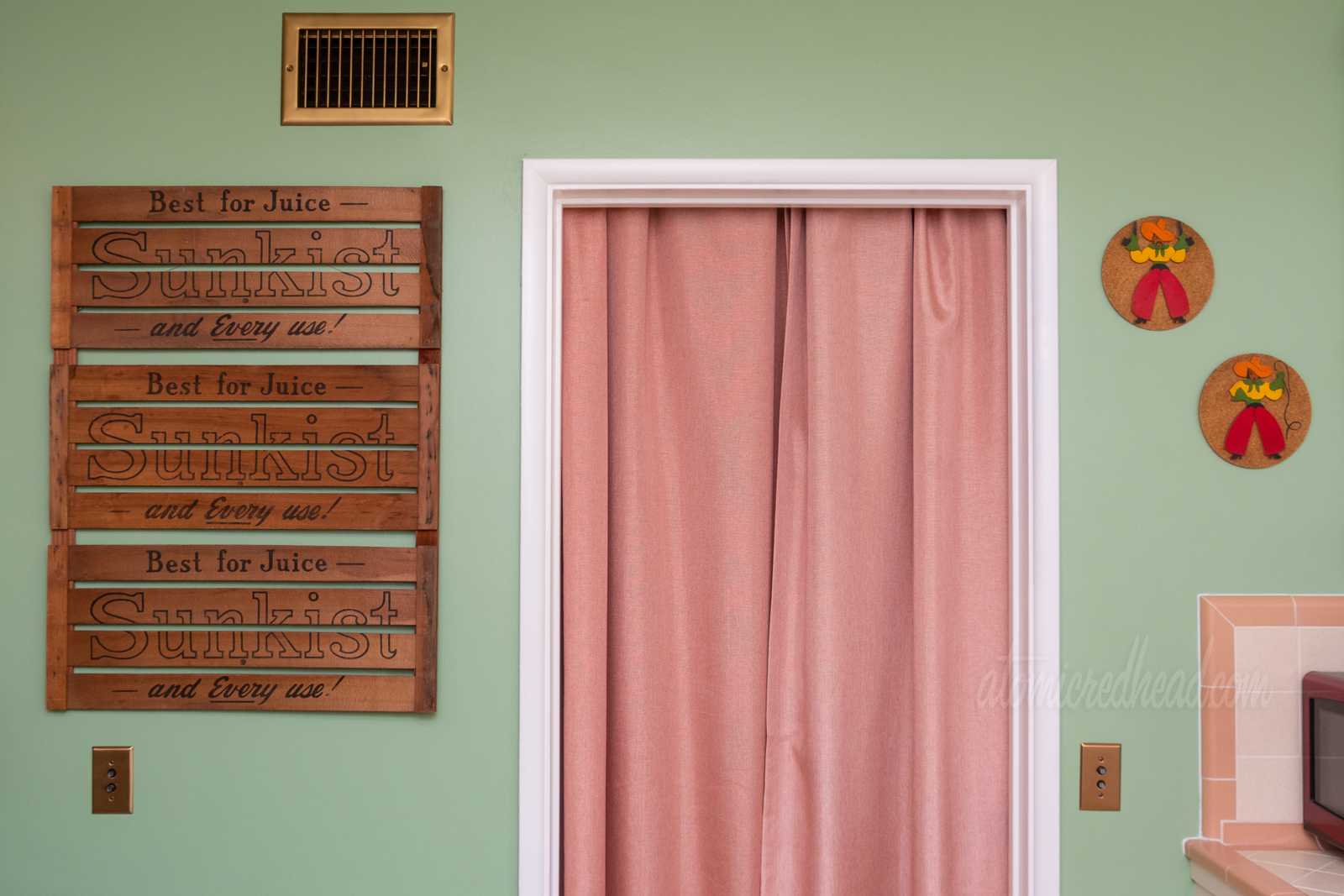 Three wood crate sides hang on a green wall to the left of a doorway that has a pink curtain. Text on the crates reads "Sunkist Best for Juice - and Every Use"