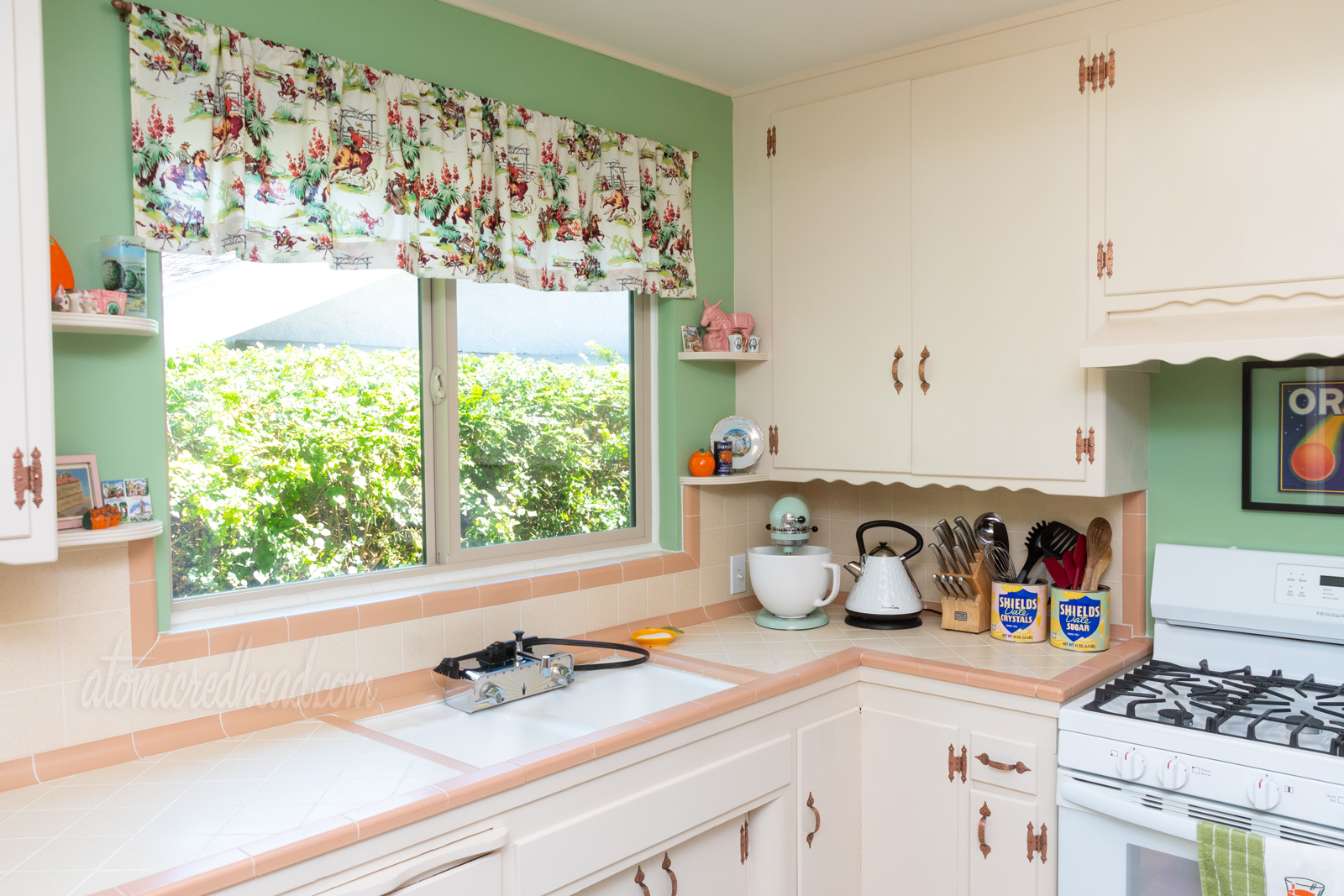 View toward the sink, which sits under a window. The window has a valance featuring cowboys near a campfire, roping steer, and yucca plants. The color scheme of the fabric is pink, green, and brown. A green Kitchenaid mixer sits in the corner.