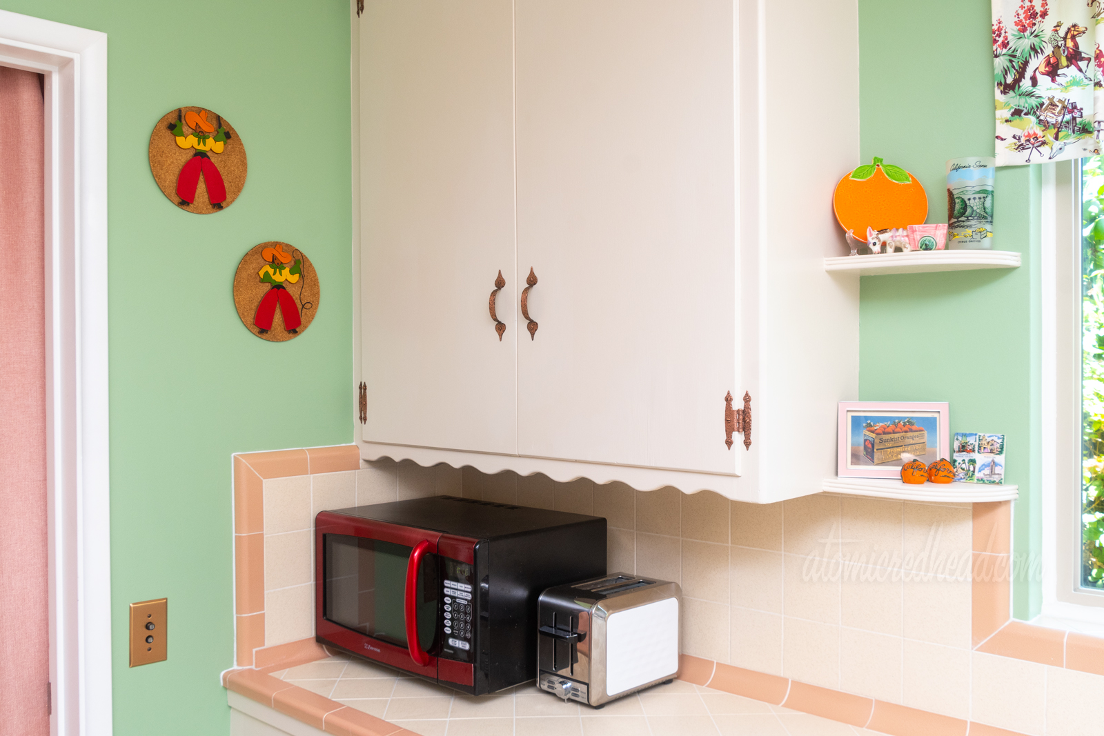 A red microwave rests on cream and pink tile counter. To the left and above the microwave are two circular pieces of art, cut wood cowboy and cowgirl that are mounted on cork.