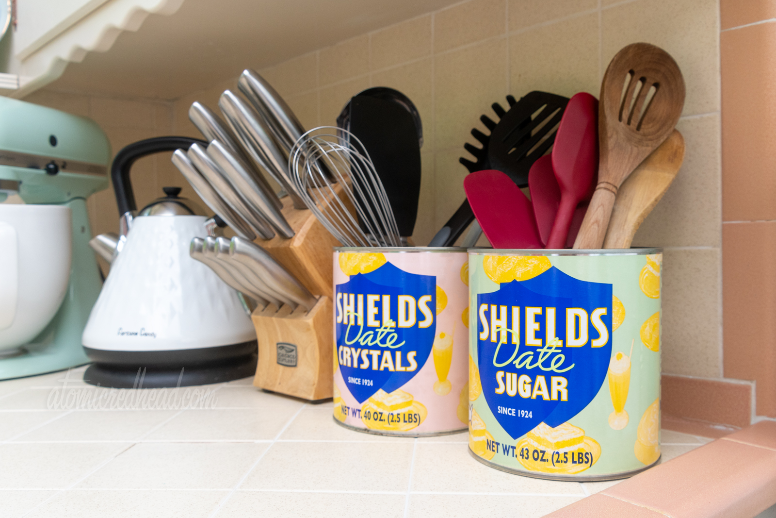 Two mental cans, one for Shields Date Sugar, the other for Shields Date Crystals, one pink and one green, sit on our counter, they house various kitchen utensils.