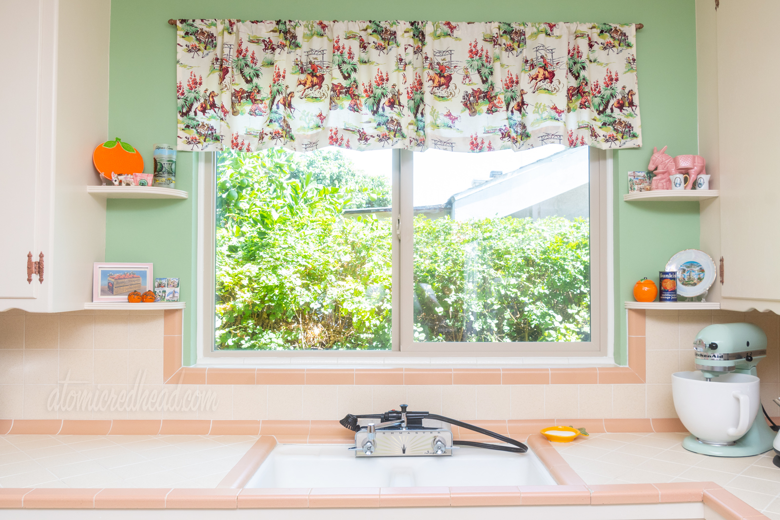 Direct view of sink. Outside the window is a green hedge. Above the window is a cream valance with images of cowboys roping steer, riding horses, and around a campfire. The fabric design is made up of pinks, browns, and greens, similar in shade to the walls.