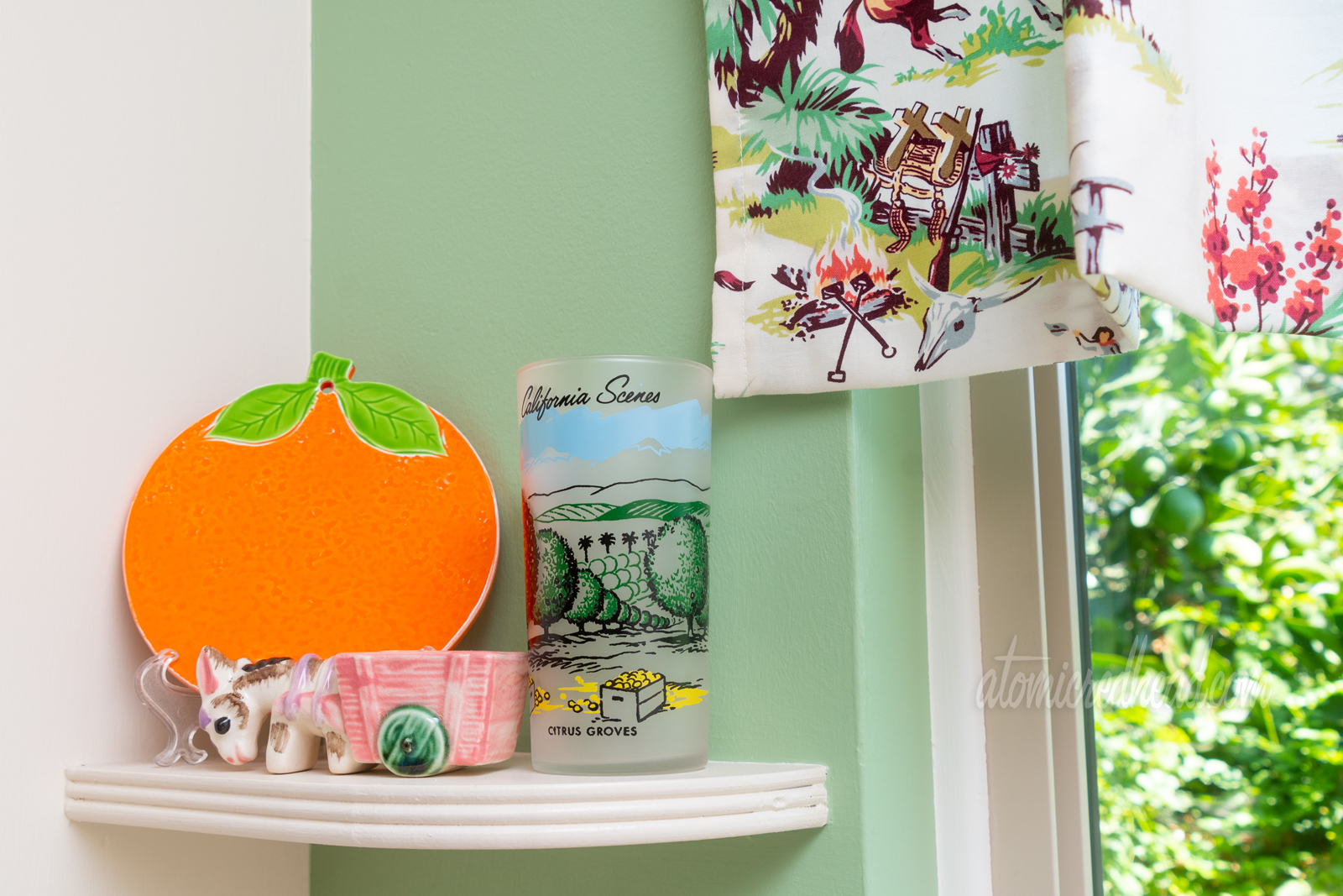 Close-up of one shelf near the sink. An orange shaped trivet leans in the corner, a glass sits next to it featuring a scene of an orange grove, and a small planter of a donkey pulling a cart.