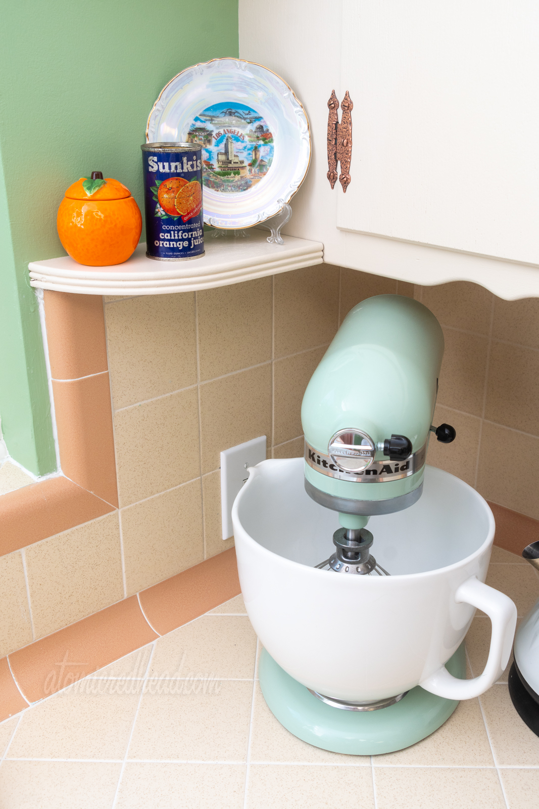A look at another shelf near the sink, on it sits a small orange shaped container, a vintage can for Sunkist concentrated California orange juice, and a souvenir plate of LA, featuring LAX, Chinatown, Griffith Observatory, Farmer's Market, and Olvera Street.