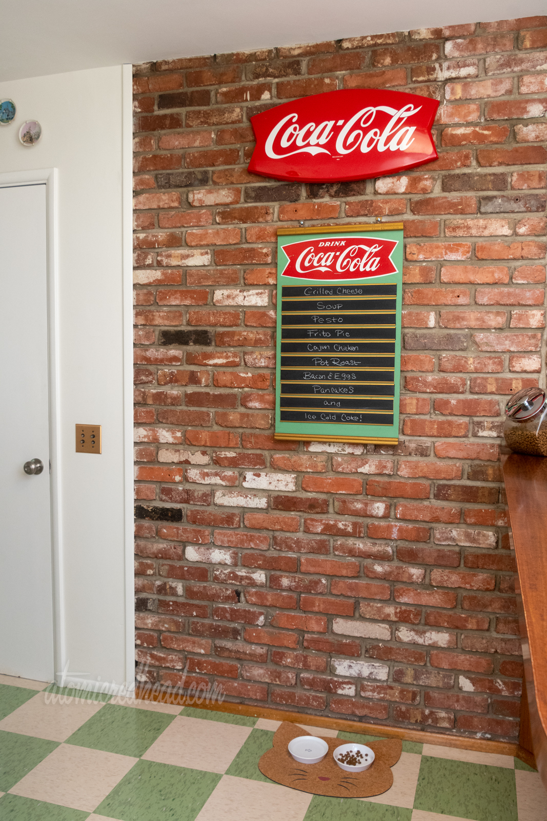 Hanging on a brick wall are a large fishtail Coca-Cola sign, and a fishtail Coca-Cola menu board which has various menu options written on chalkboard slats. Under the menu, on the floor sits a cork mat shaped like a cat head, where the eyes are are two bowls, one for water, the other for food. The floor is checkerboard of cream fleck and green fleck tiles.