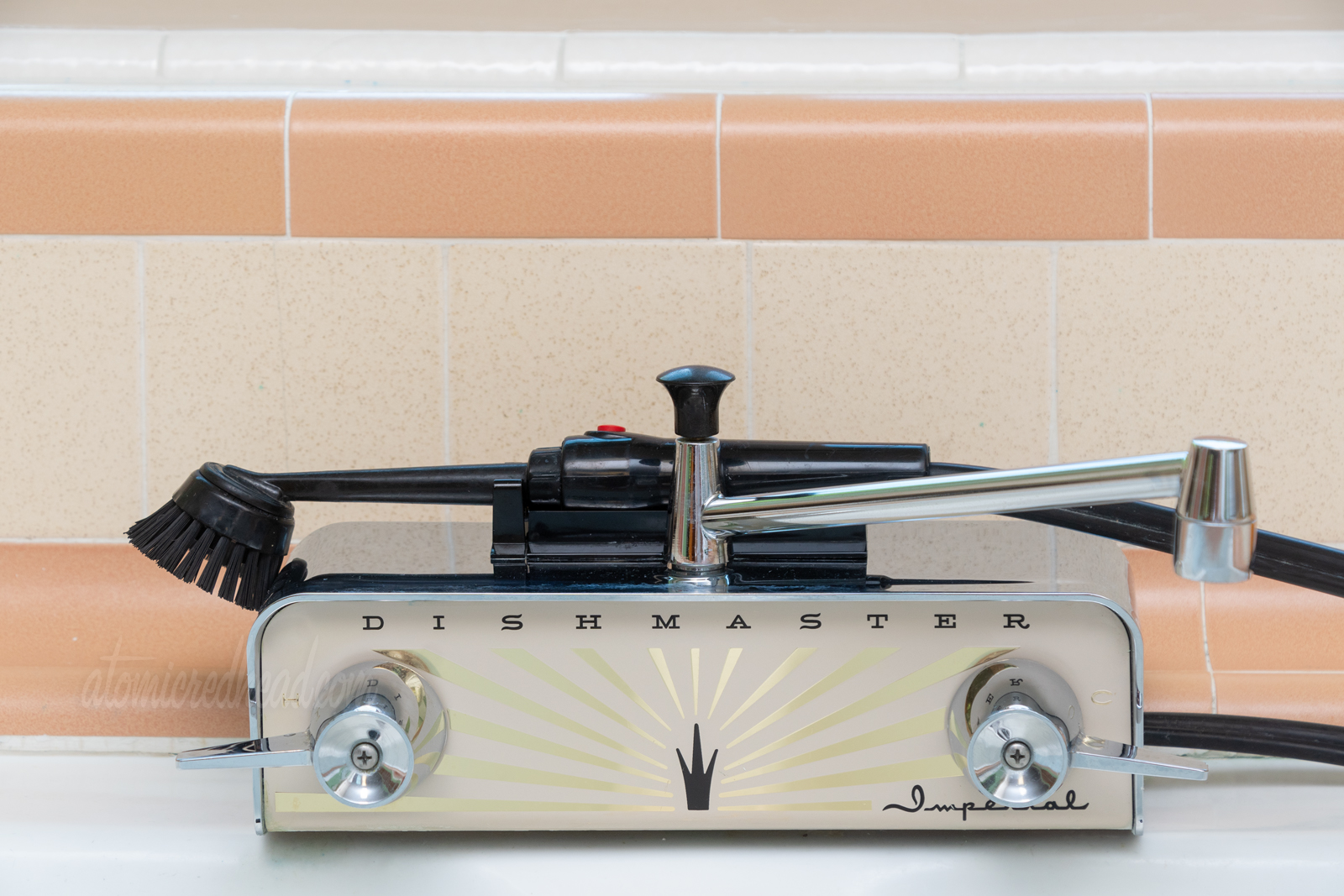 Close-up of our facet, a chrome rectangle features a faceplate of cream, with a gold burst coming from a small black crown. Black text reads "Dishmaster" across the top. 