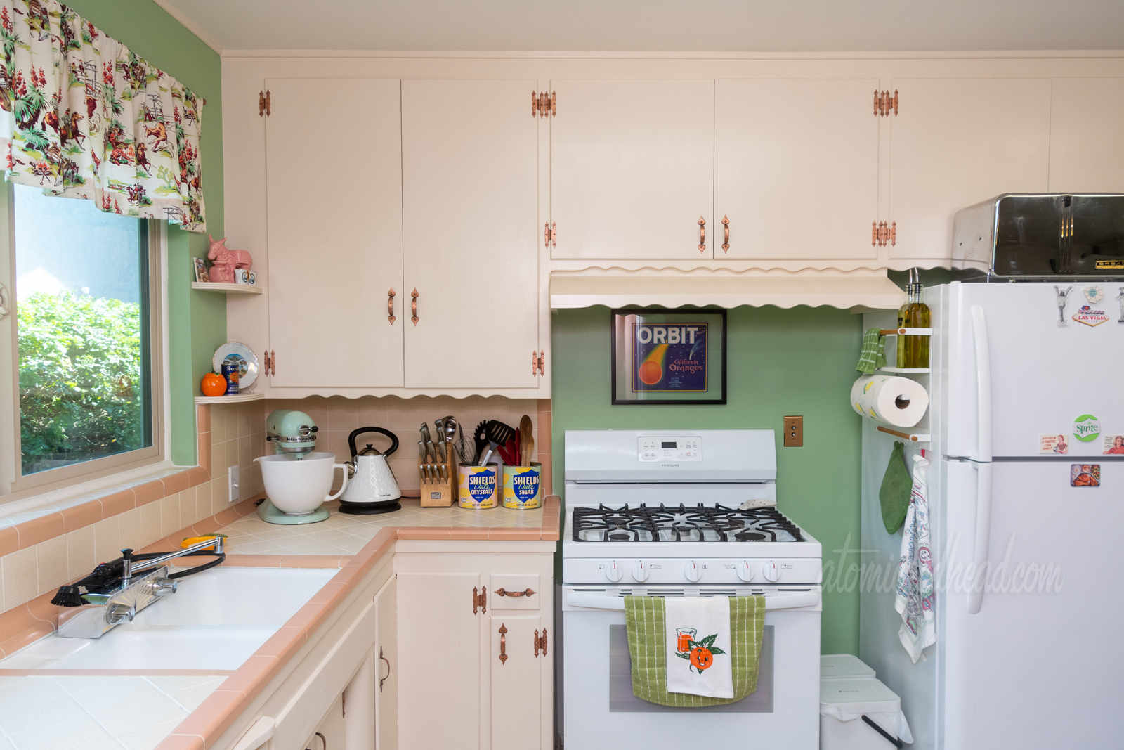 A view toward the stove, which is tucked under cream cabinets and against a green wall. Above the stove hangs a vintage fruit crate label that has an orange streaking across the sky like a comet, and above it reads "Orbit" in large white letters. Hanging on the handle of the stove is a white towel with an orange with a face o it.