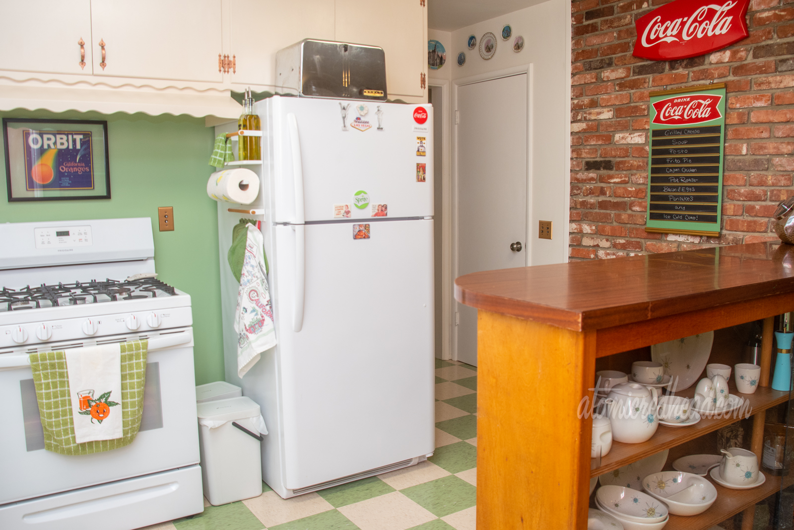 A white stove sits to the left of the fridge, just pas the fridge is a hallway with a doorway, next to the door a brick wall that has a Coca-Cola menu hanging on it. A bar with built in china hutch juts out from the right side.