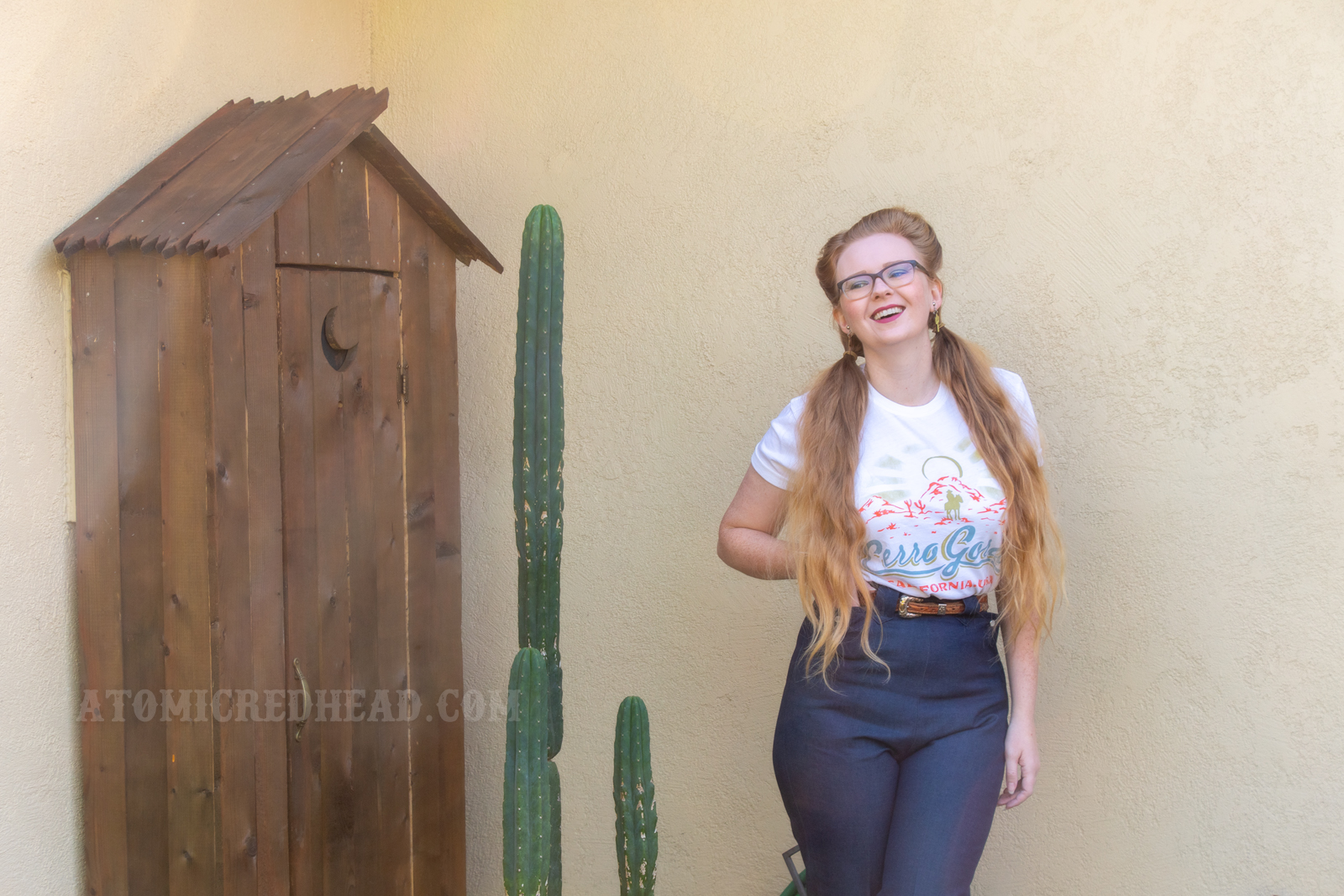 Myself standing in front of a western style outhouse with cacti in front, wearing a Cerro Gordo tee and western style jeans.