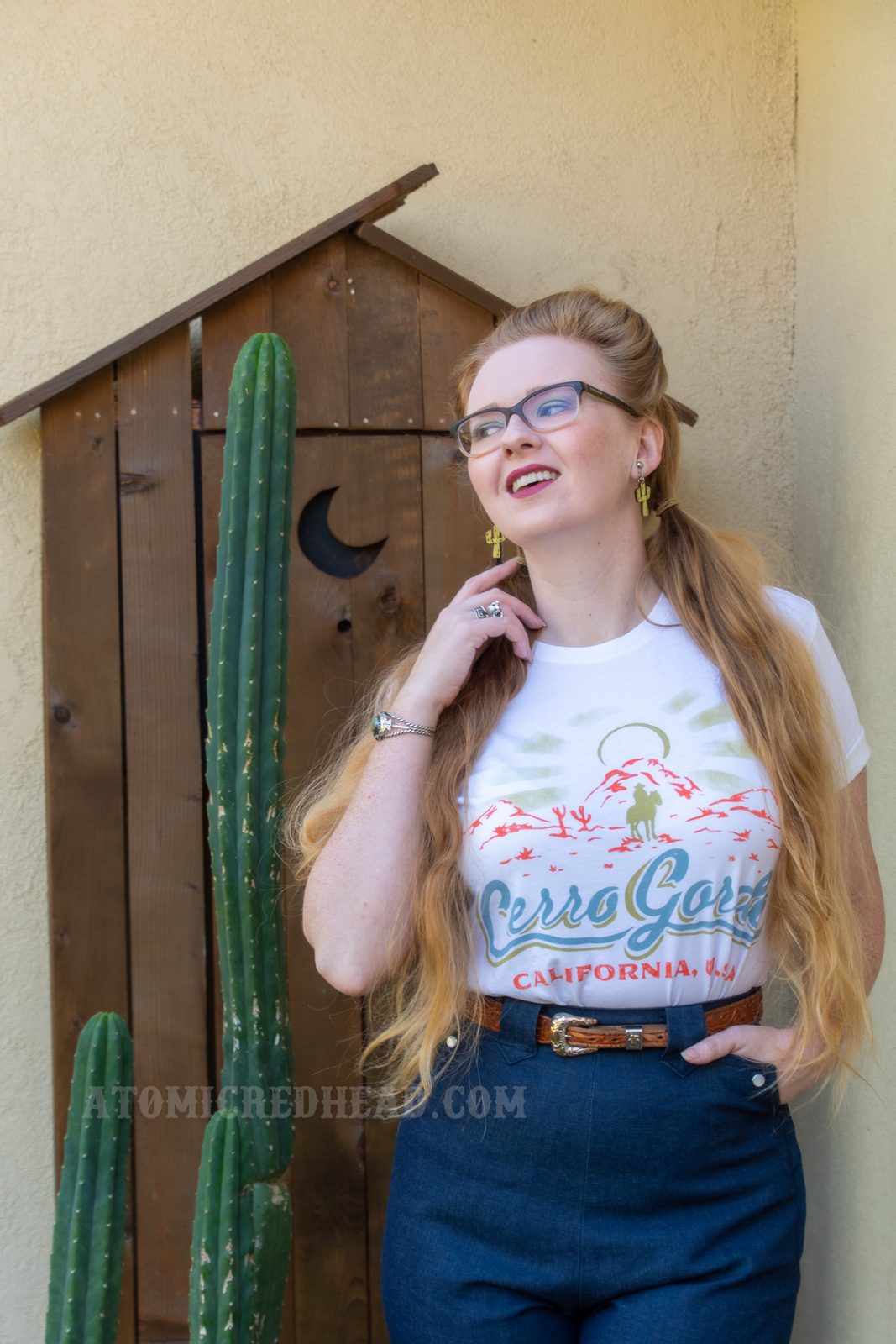 Myself standing in front of a western style outhouse with cacti in front, wearing a Cerro Gordo tee and western style jeans.