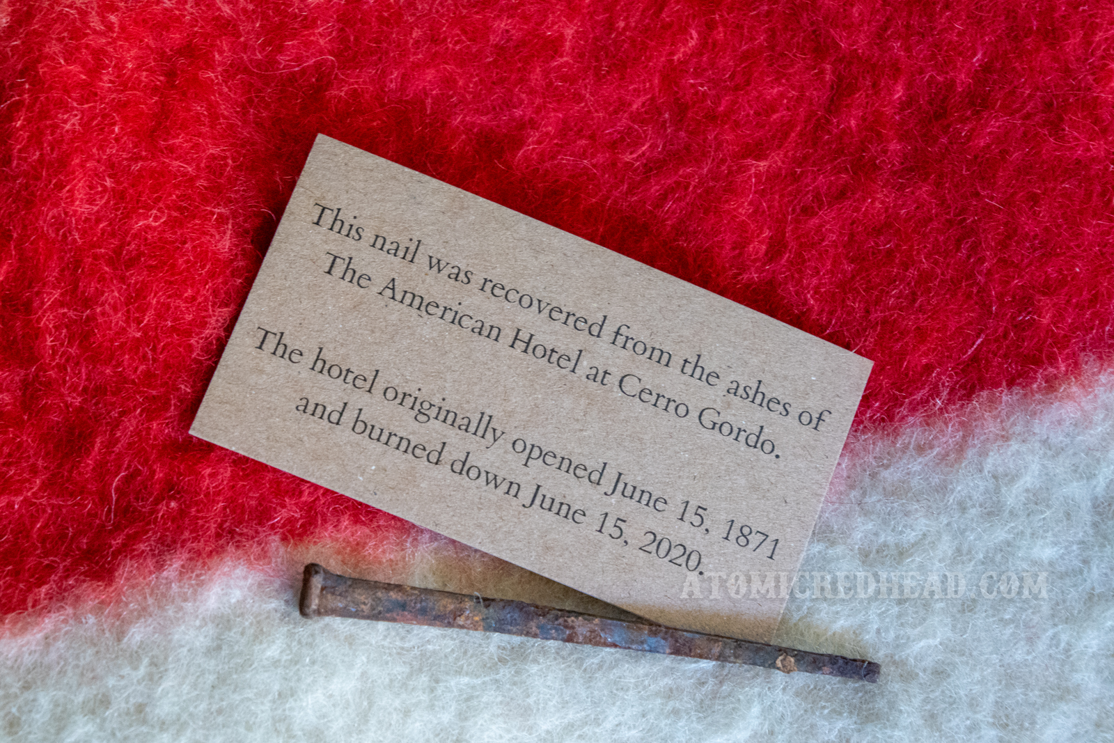A rusty square head nail next to a card that reads "This nail was recovered from the ashes of The American Hotel at Cerro Gordo. The hotel originally opened June 15, 1871 and burned down June 15, 2020."