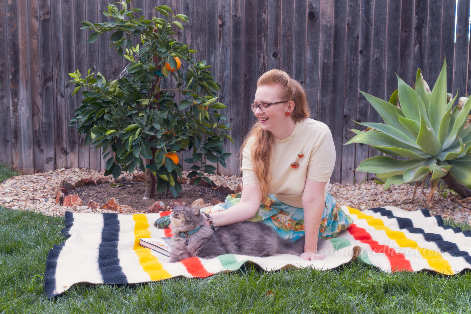 Sitting on a blanket in front of my orange tree, wearing a white sweater, with two orange brooches, and a skirt featuring various California landmarks, while reading and petting my cat.