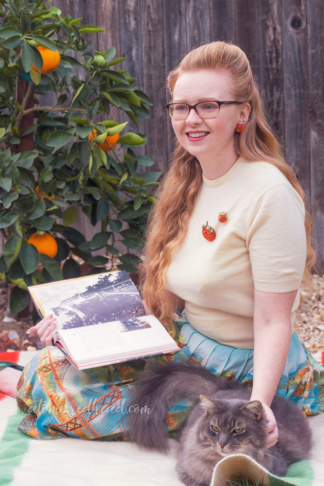 Sitting on a blanket in front of my orange tree, wearing a white sweater, with two orange brooches, and a skirt featuring various California landmarks, while reading and petting my cat.