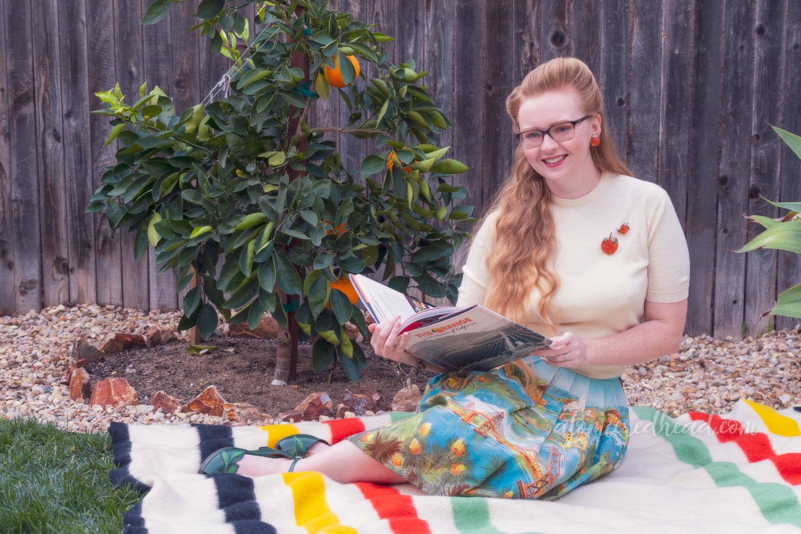Sitting on a blanket in front of my orange tree, wearing a white sweater, with two orange brooches, and a skirt featuring various California landmarks, while reading.