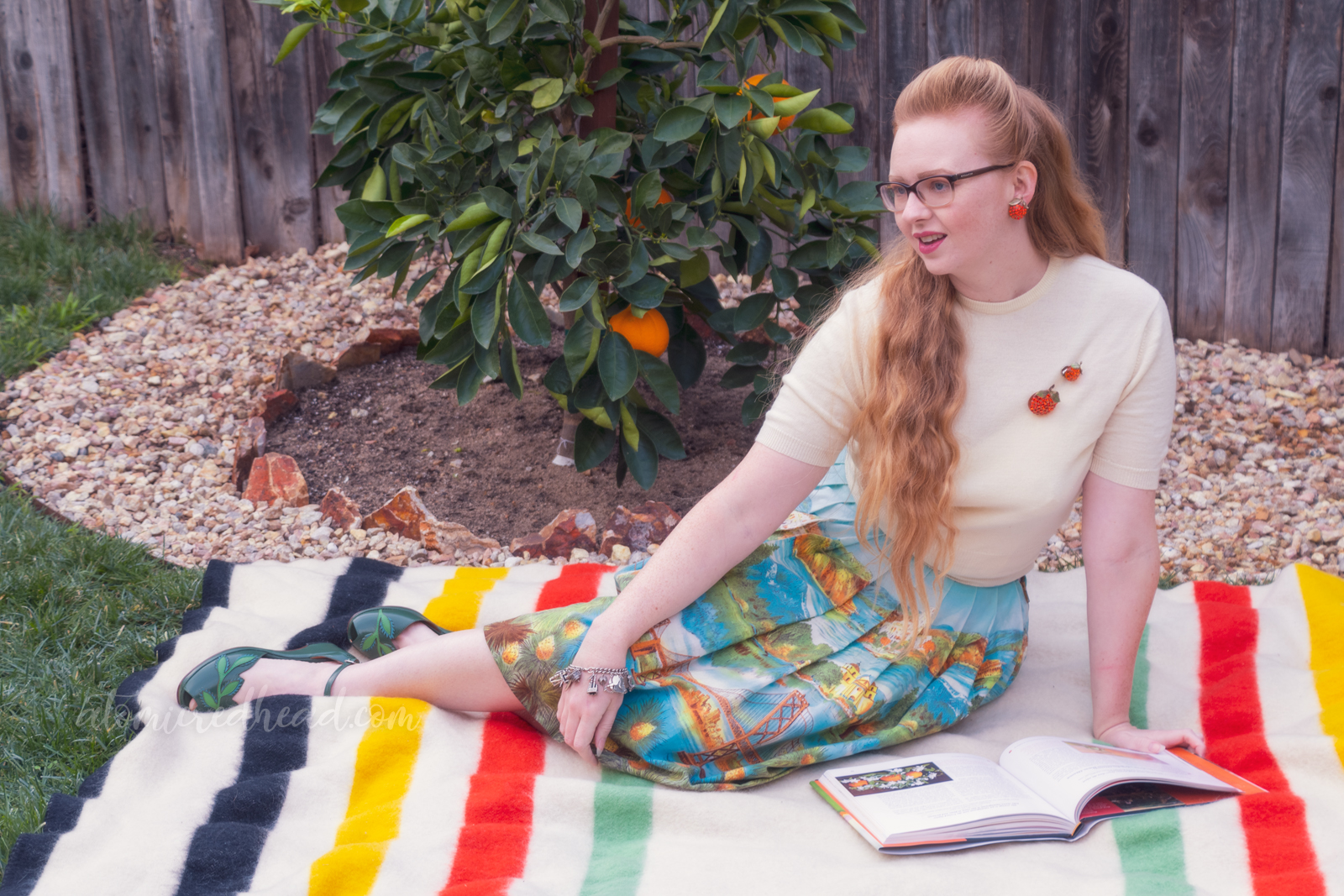Sitting on a blanket in front of my orange tree, wearing a white sweater, with two orange brooches, and a skirt featuring various California landmarks, while reading.