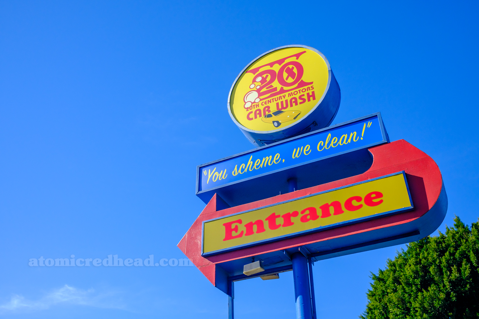 A large, red, blue, and yellow sign reading "20th Century Motors Car Wash 'You Scheme, We Clean,' Entrance" With the "Entrance" in a red arrow.