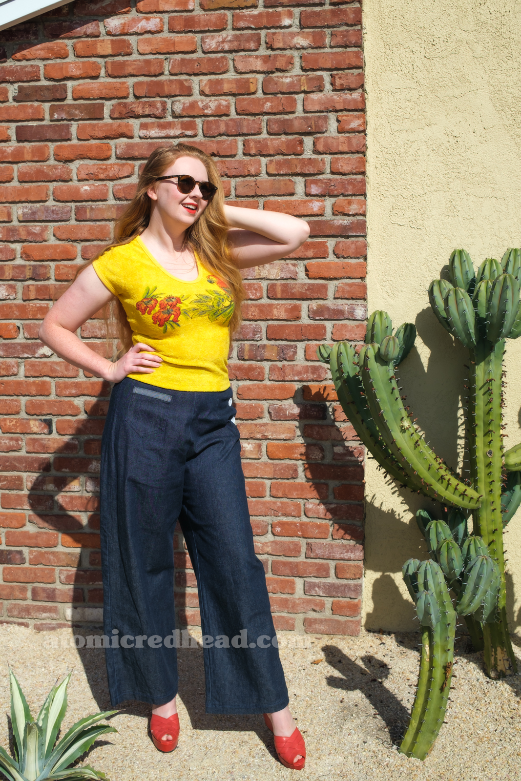 Myself, wearing a yellow terrycloth shirt featuring red hibiscus flowers and a parrot, blue wide leg jeans, red platform shoes, standing outside against a brick wall.