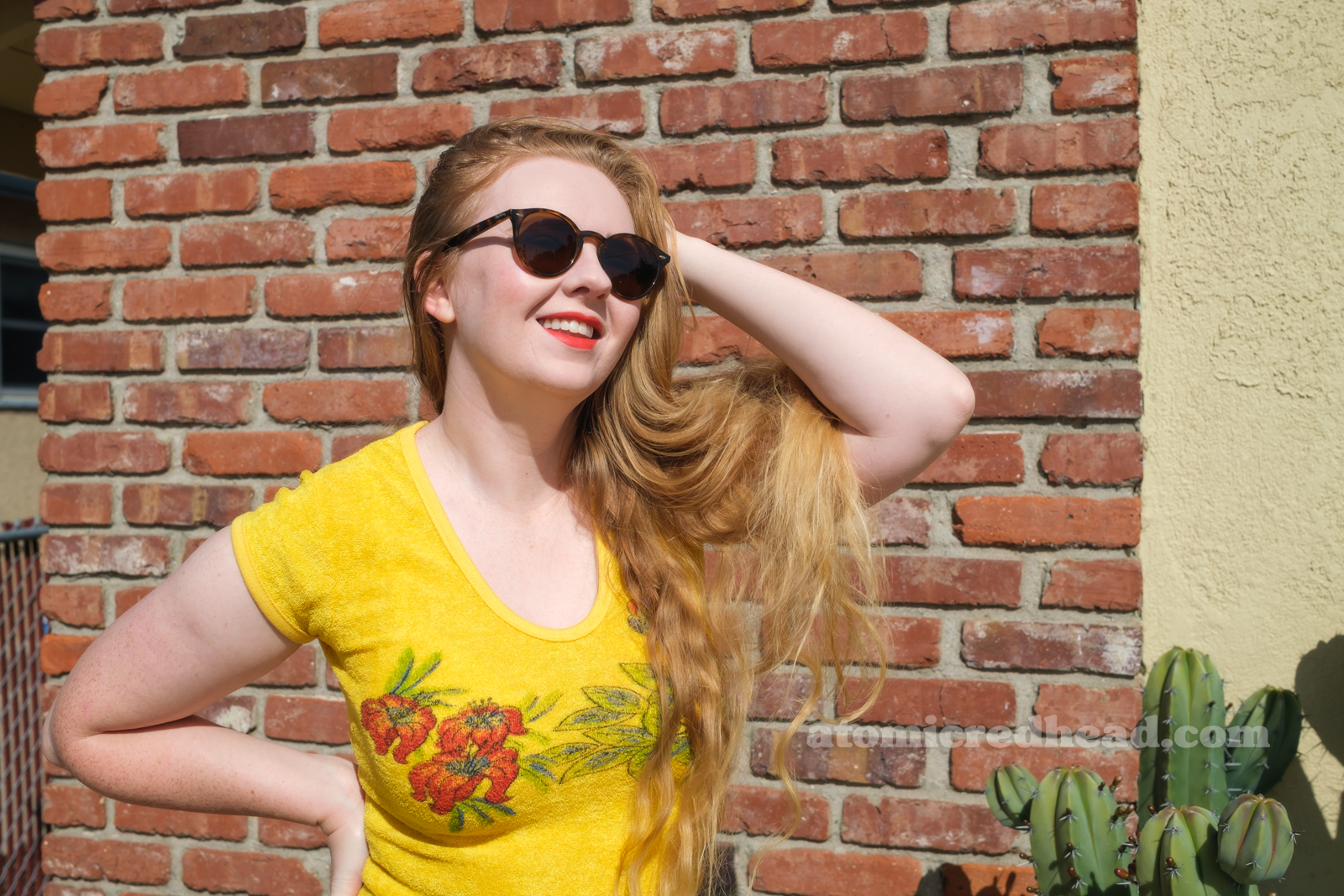 Myself, wearing a yellow terrycloth shirt featuring red hibiscus flowers and a parrot, blue wide leg jeans, red platform shoes, standing outside against a brick wall.