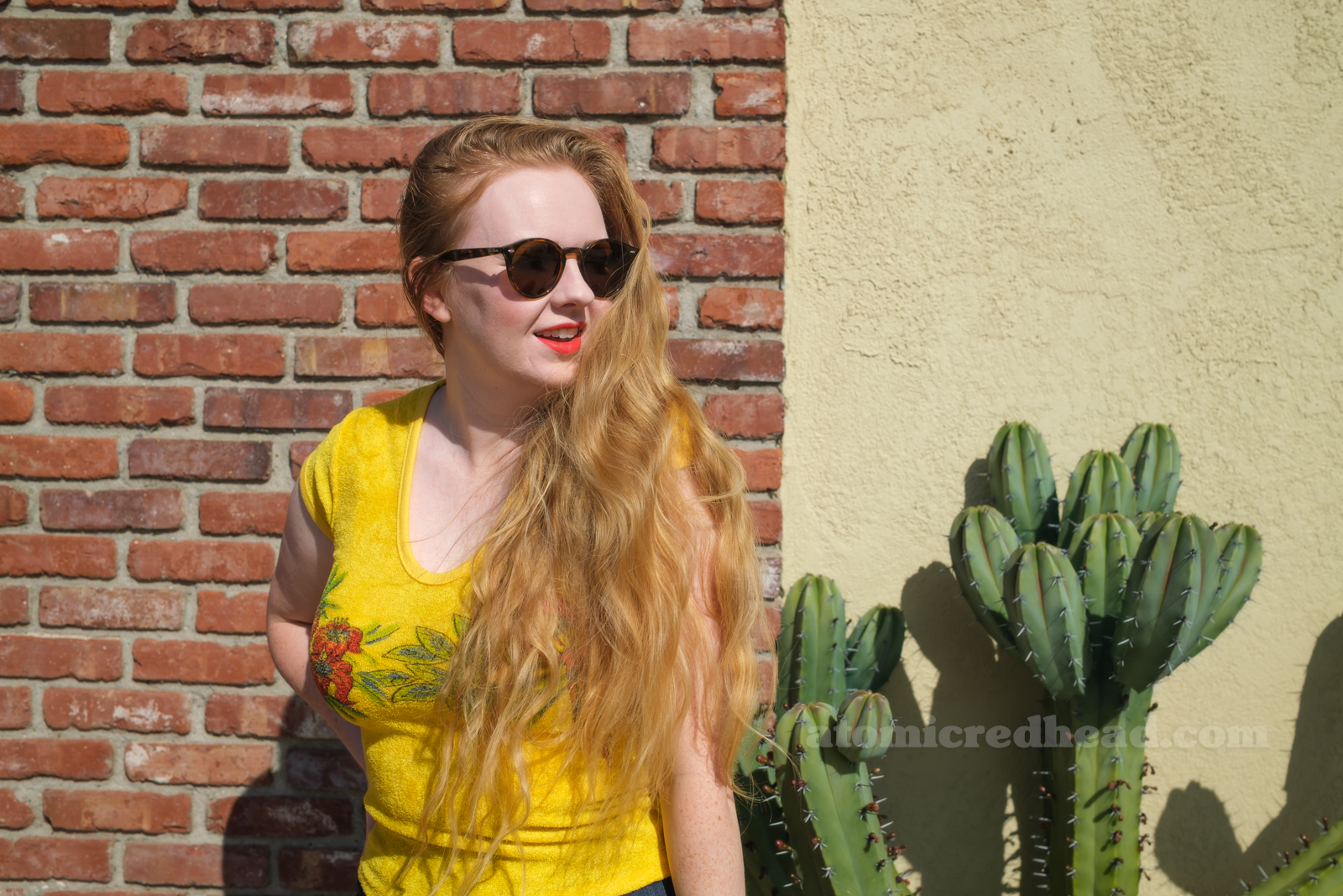 Myself, wearing a yellow terrycloth shirt featuring red hibiscus flowers and a parrot, blue wide leg jeans, red platform shoes, standing outside against a brick wall.