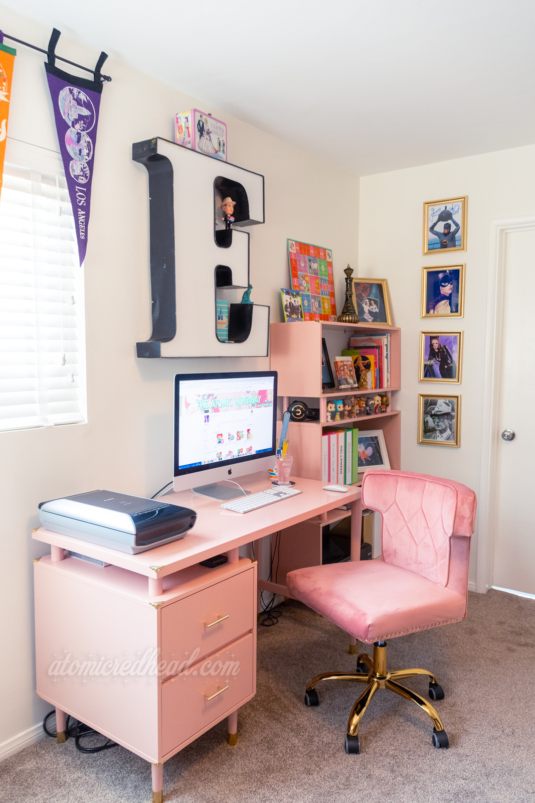 Overview of my corner. A computer sits atop a pink desk, with a pink velvet chair at it, behind it is a matching pink bookcase. A giant letter E hangs above the computer.