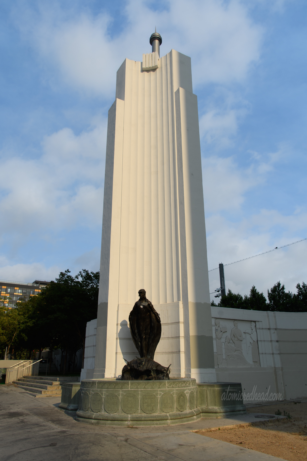 A tall pillar that is curved and done in the Art Deco style, features a statue of a woman who rides a small wave with flying fish emerging from it at the base.