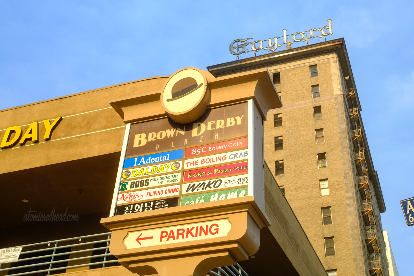 Sign for Brown Derby Plaza. At the top is a small image of a brown bowler hat and text below reads "Brown Derby Plaza" signs for other businesses are below, and include a bakery, dentist, and Korean food. 