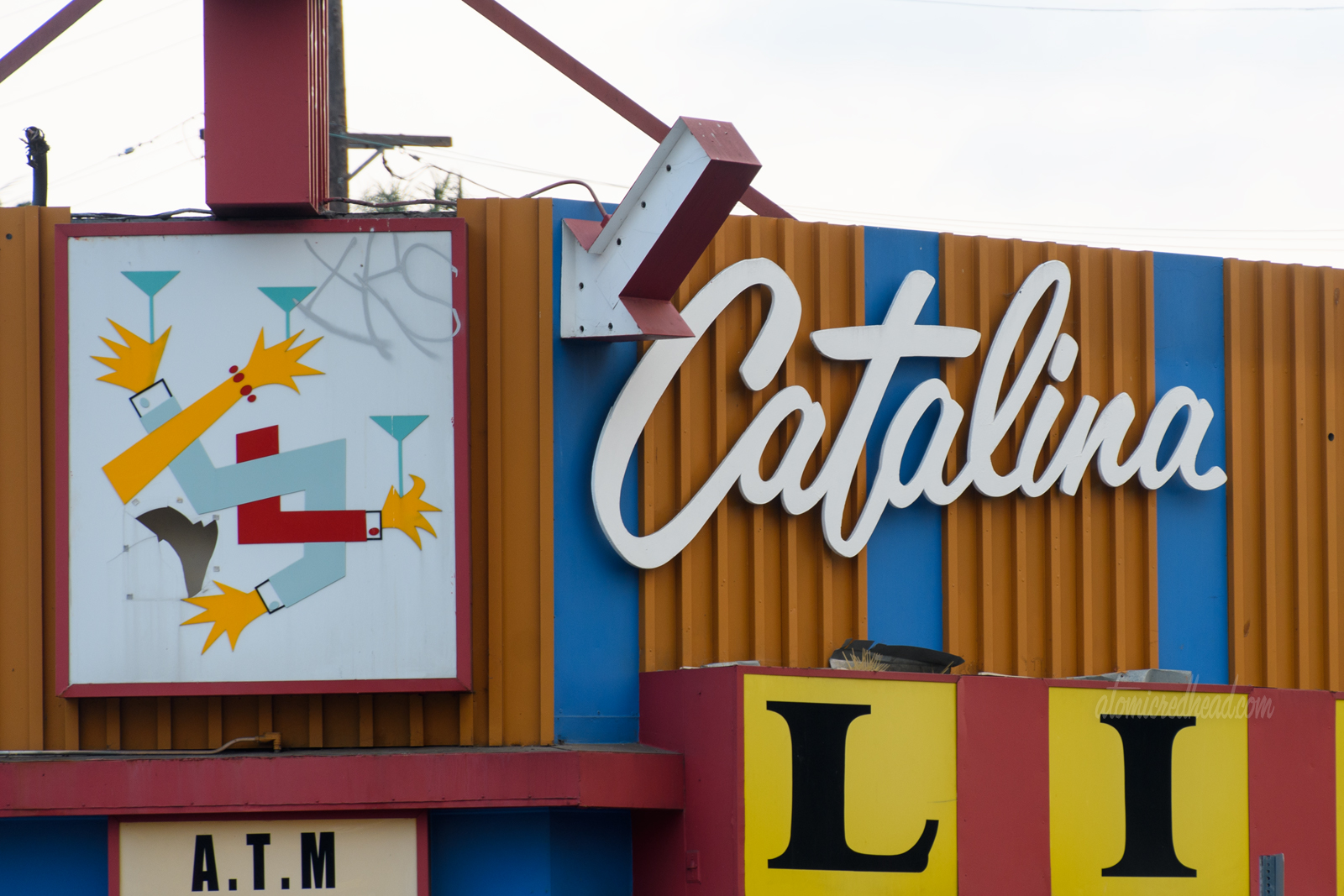 The signage for Catalina Liquor, all of which is backlit plastic. Above the door is a sign that is an abstract of various arms intertwined holding martini glasses. 