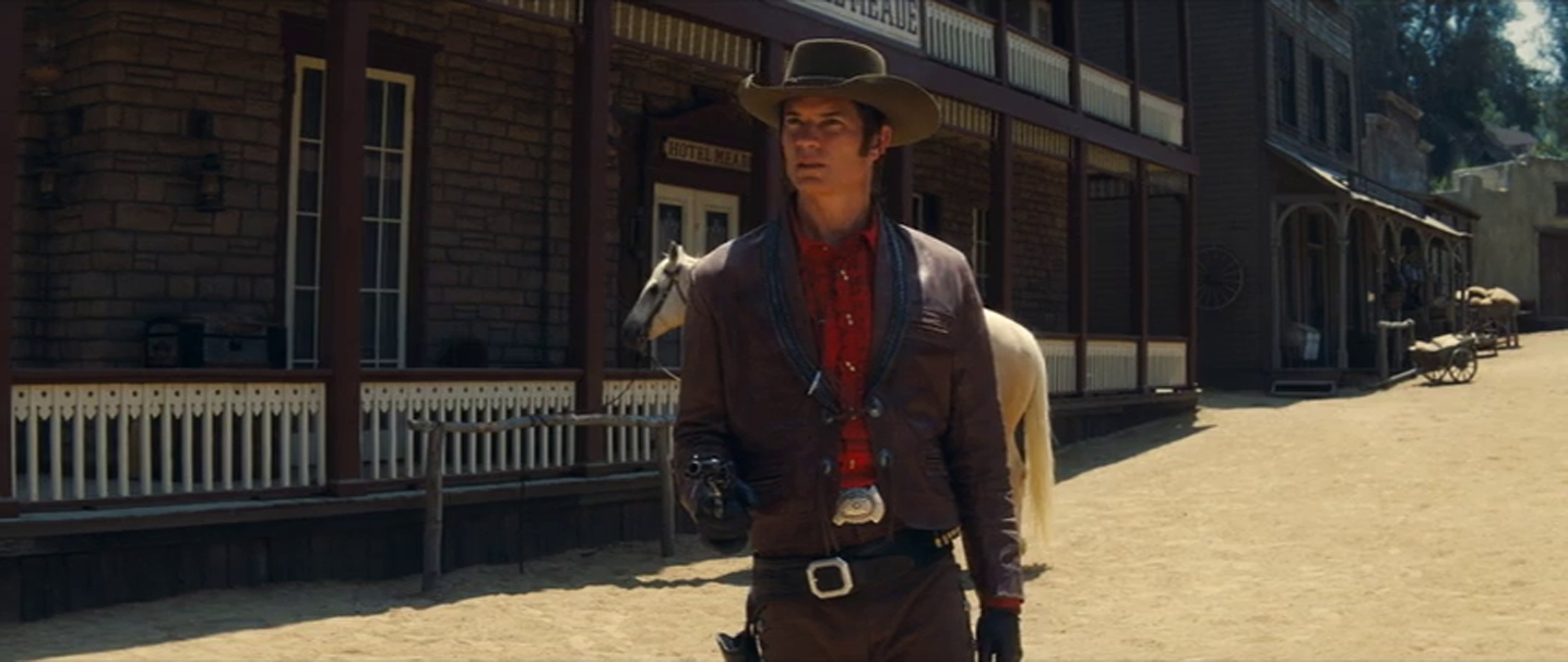 Timothy Olyphant, dressed as a cowboy films a scene for Lancer, a grey stone building with maroon trim is behind him.