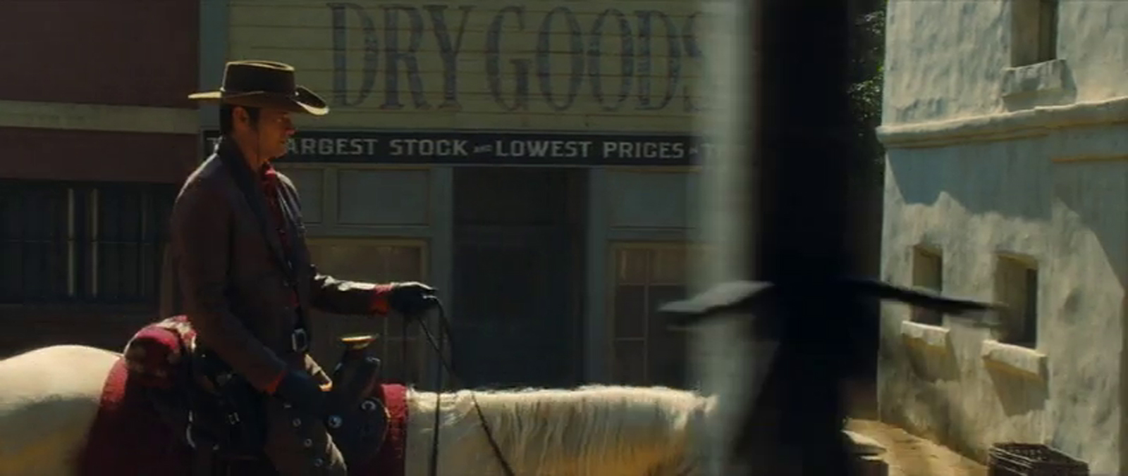 Timothy Olyphant rides a horse into the scene, passing by a wooden store with "Dry Goods" painted on the front.