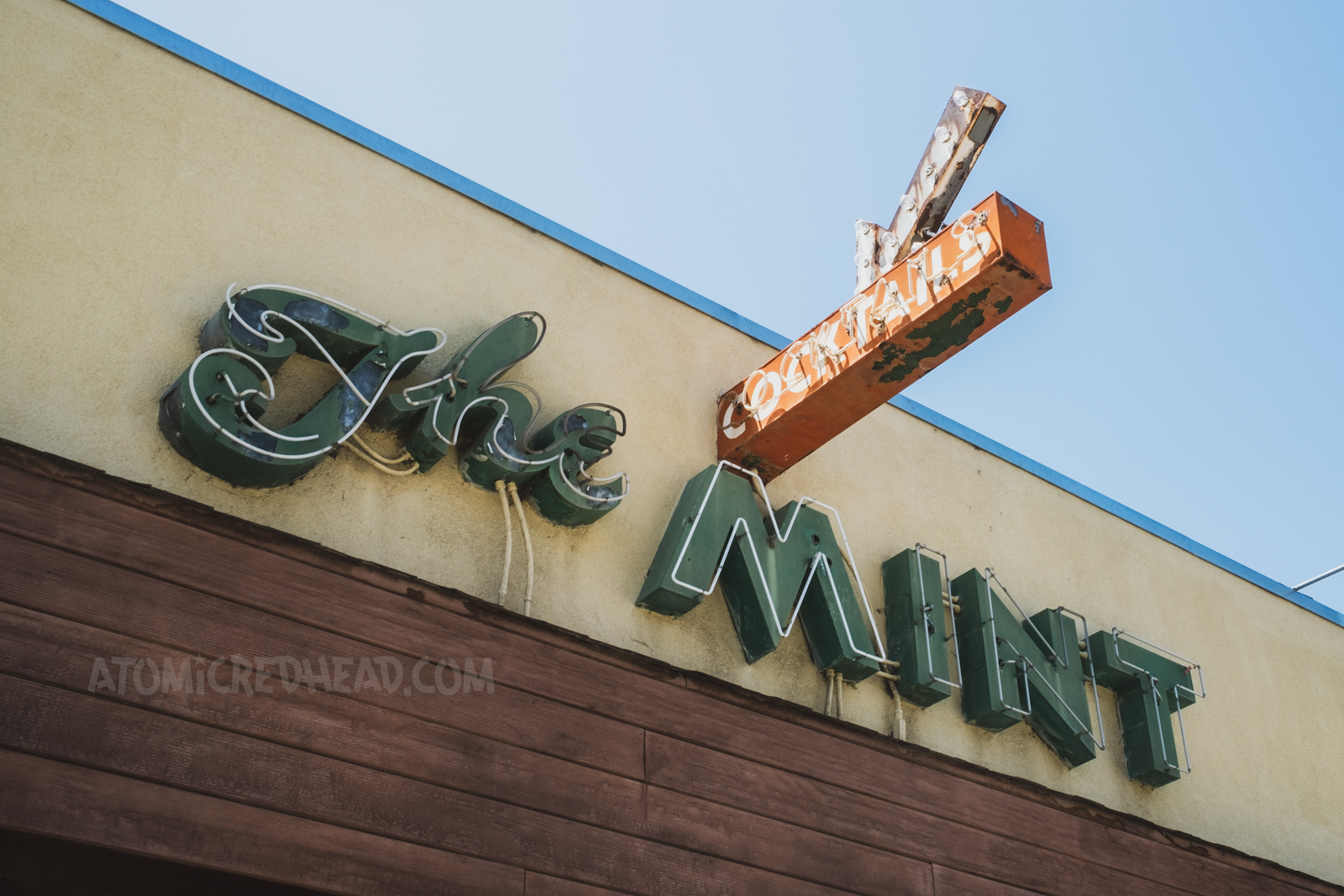 Green neon letters reading "The Mint" are attached to the side of a stucco building. A small red sign juts out from the building reading "Cocktails" and features a small arrow above.