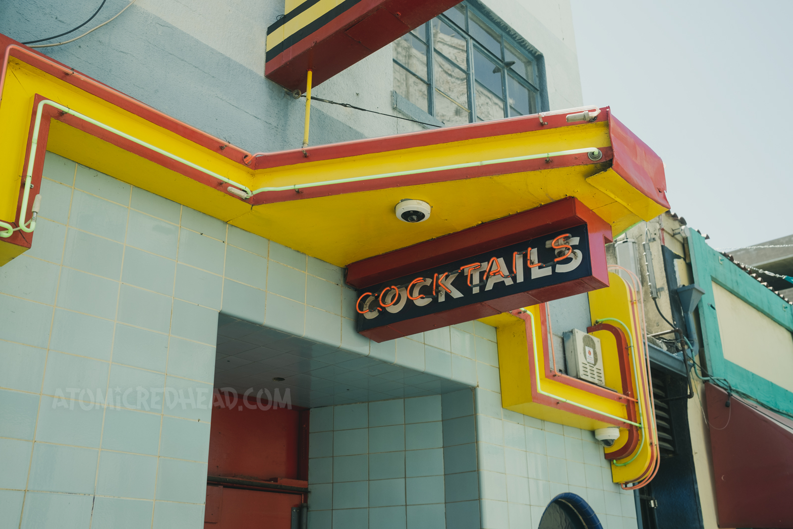 An Art Deco style building of blue tile features a small yellow awning with a neon "Cocktails" sign hanging from it.